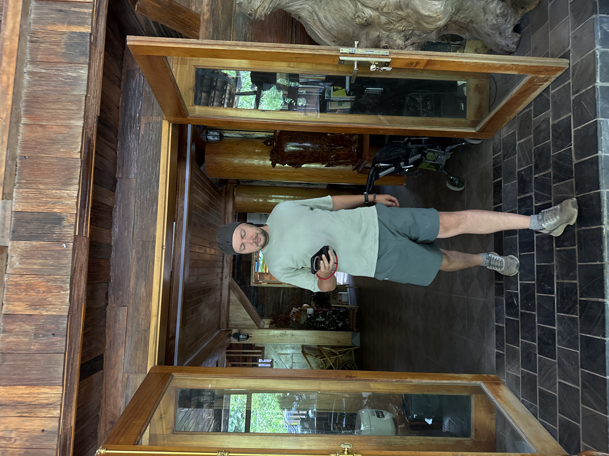 In Bản Đêu, Vietnam, a man walks forward from the interior of a wooden building, approaching the threshold defined by two open, wooden-framed glass doors. He wears a light green t-shirt, dark gray shorts, a dark gray beanie, and gray athletic shoes, with a red wristband on his left wrist. He is actively looking at a black smartphone held in his left hand.
The building's interior features extensive dark and light brown wooden plank walls and ceilings. The floor at the immediate entrance consists of dark rectangular tiles laid in a brick pattern. Beyond the man, the interior reveals various furnishings including a black baby stroller with green elements, a large, intricately carved dark wooden urn on a pedestal, and several tall, smooth, cylindrical wooden columns. A wall-mounted lantern is visible on a wooden post. To the far right, a large, gnarled piece of natural wood stands as a decorative element.
Through the left open door, dense green foliage is visible outside, along with a white rice cooker and a distant wooden staircase. Through the right open door, shelves containing books and other items are discernible, along with more exterior greenery. The right door has a silver-colored handle with keys attached. The overall scene suggests a rustic yet well-appointed indoor environment with a strong natural aesthetic.