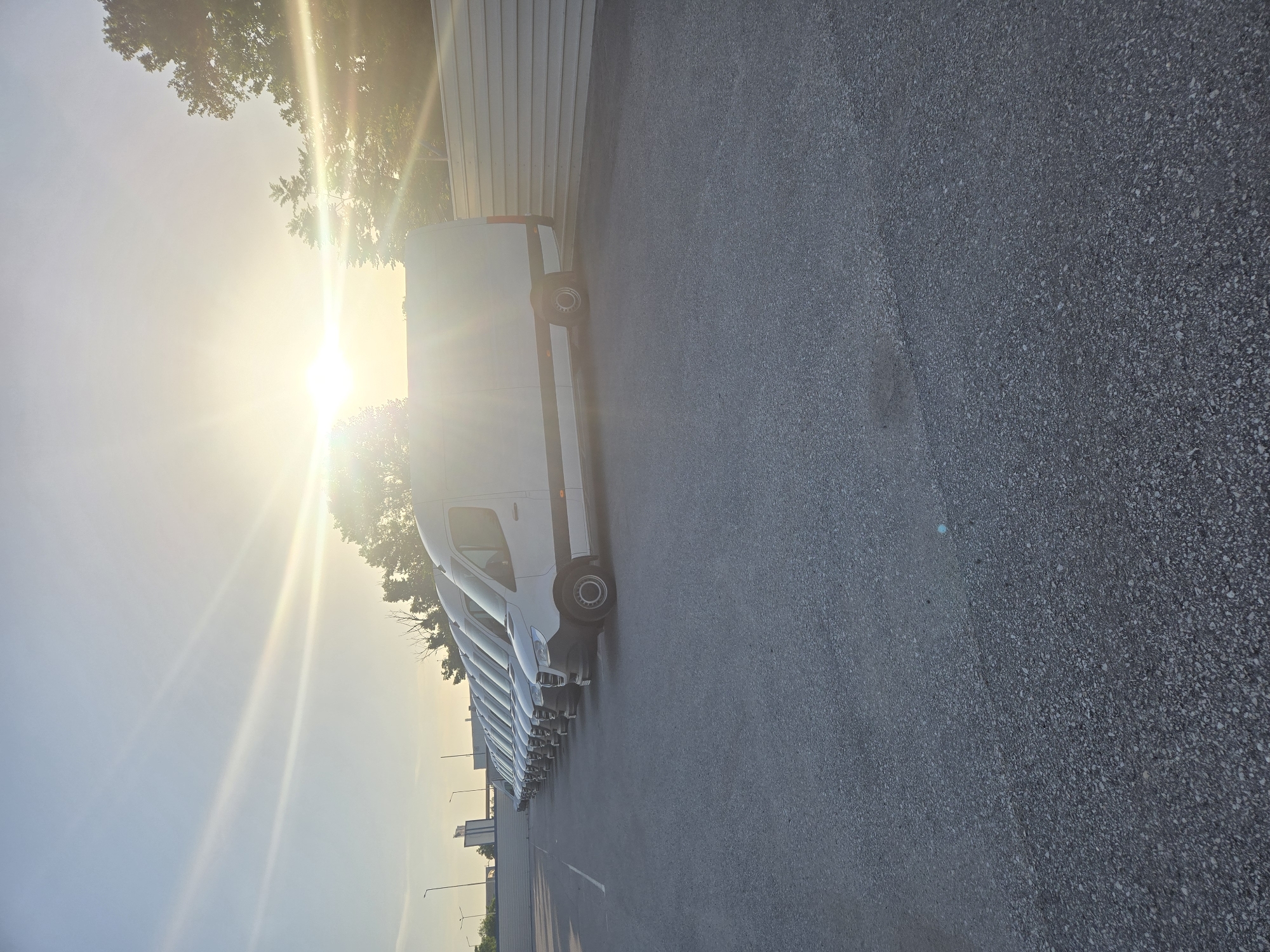 A row of white vans is parked next to a building with white siding in Unknown, Unknown. The sun is shining brightly from the upper left corner of the image. A black asphalt surface is visible in the foreground. The sky is pale blue. There are some power poles and trees in the distance.