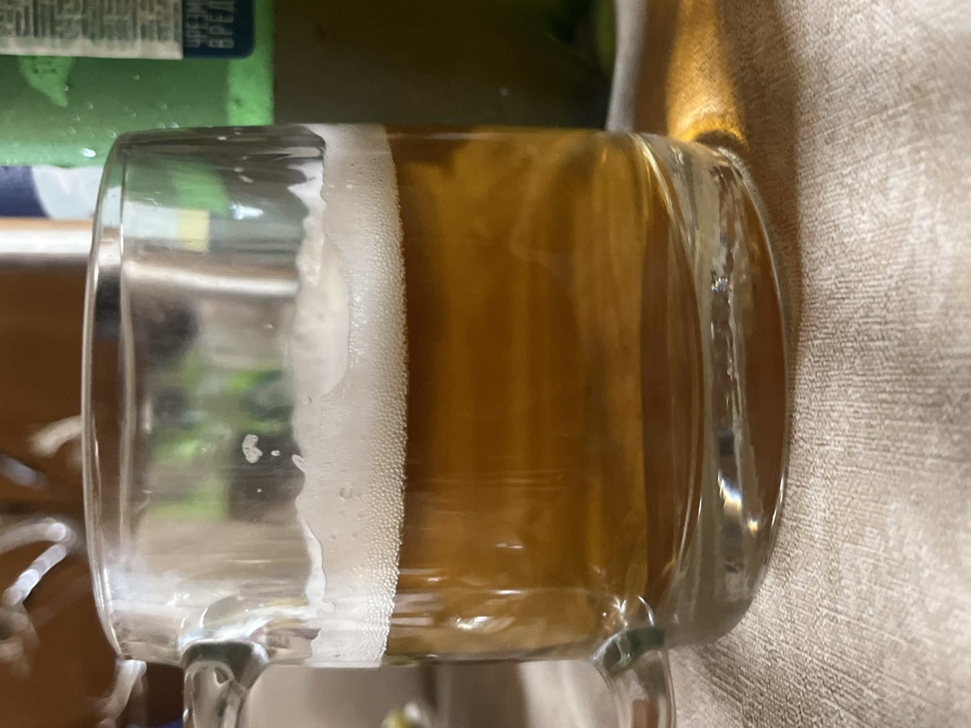 A close-up, eye-level shot shows a glass mug filled with amber-colored liquid and a frothy head. The mug has a clear handle and is positioned on a textured, light-colored surface, possibly a tablecloth. In the background, out of focus, a green bottle with Russian text is visible, suggesting the beverage might be beer. The lighting casts a warm glow, highlighting the condensation on the bottle and the reflections within the glass. The scene appears to be indoors, likely in a home or a casual dining setting, in the city of Vasilyevo, Russia. No people are visible in the image.