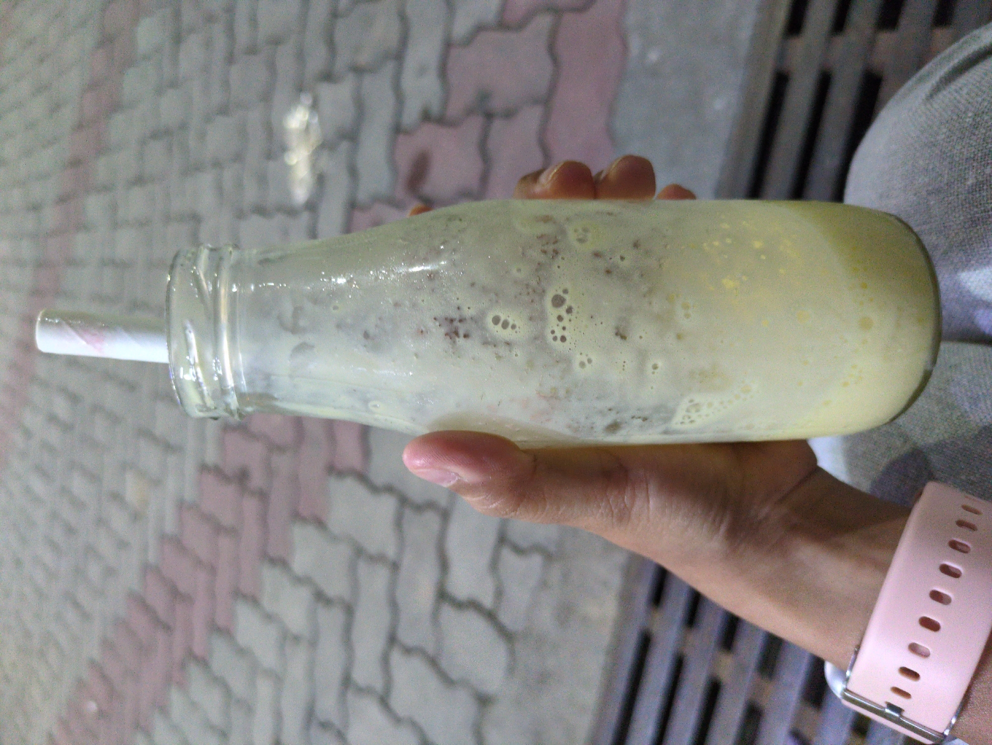 A close-up, waist-up shot shows a person's left hand holding a glass bottle filled with a frothy, pale yellow beverage and a white and pink striped straw. The bottle is partially obscured by the hand, which is positioned towards the bottom of the frame. The person is wearing a light pink, perforated watchband on their left wrist. The background consists of a patterned, brick-like pavement in shades of grey and pink, with a dark metal grate visible in the lower left corner. The scene appears to be outdoors, possibly in a public area like a cafe or street in Dharamshala, India, given the presence of the paving stones and the outdoor setting. The beverage has small air bubbles and some darker specks within it.