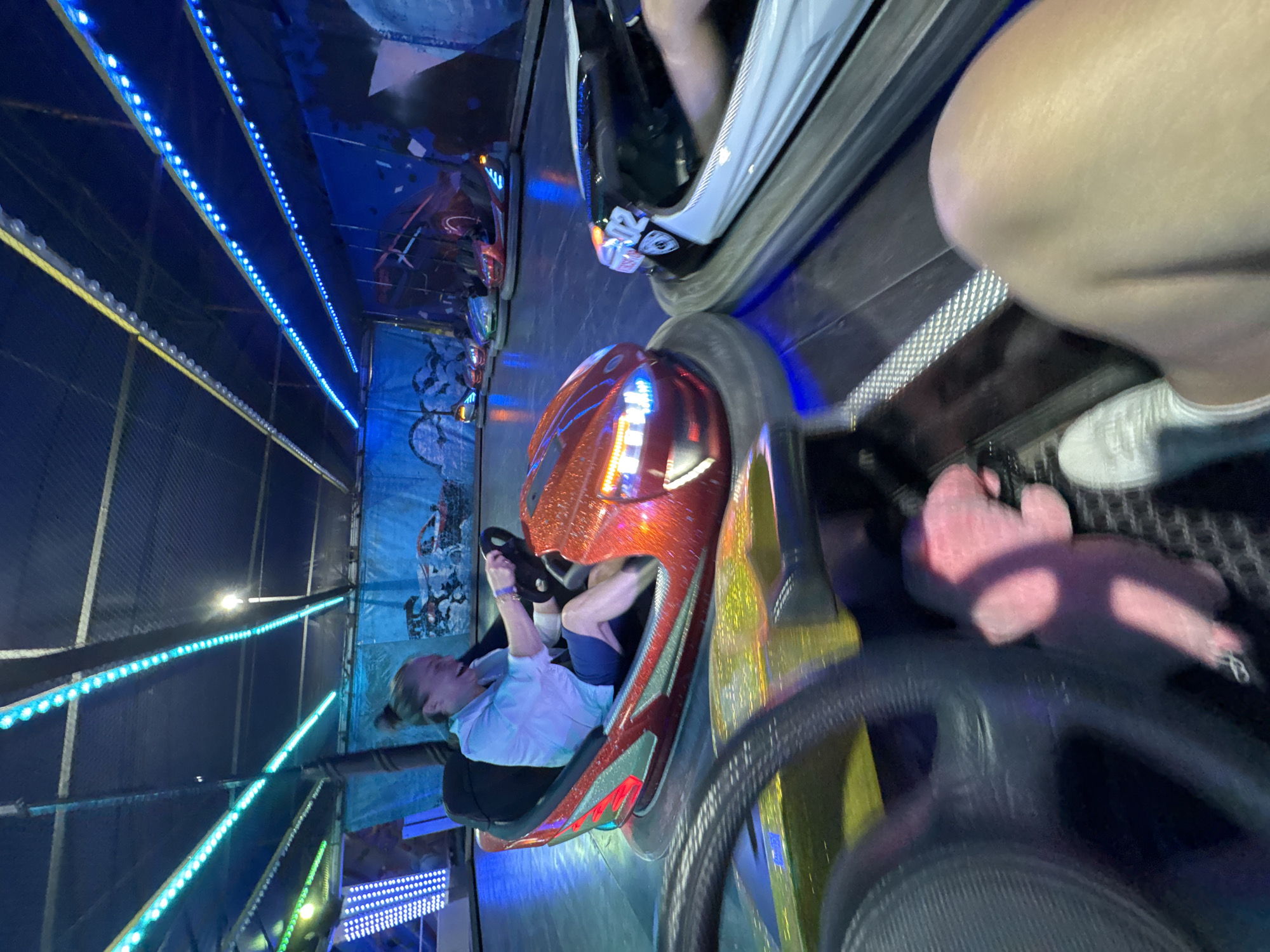In Nesebar, Bulgaria, a woman in a light blue shirt and dark shorts is driving a red bumper car, laughing. She is holding the steering wheel of the bumper car, which has sparkly detailing. The bumper car is on a track, and other bumper cars are visible in the background, some with bright LED lights. The ceiling of the arena is decorated with blue and green LED strips. The image is taken from the perspective of another bumper car, showing its steering wheel and the leg of its driver in the foreground. The overall atmosphere is lively and fun, typical of an amusement park attraction.