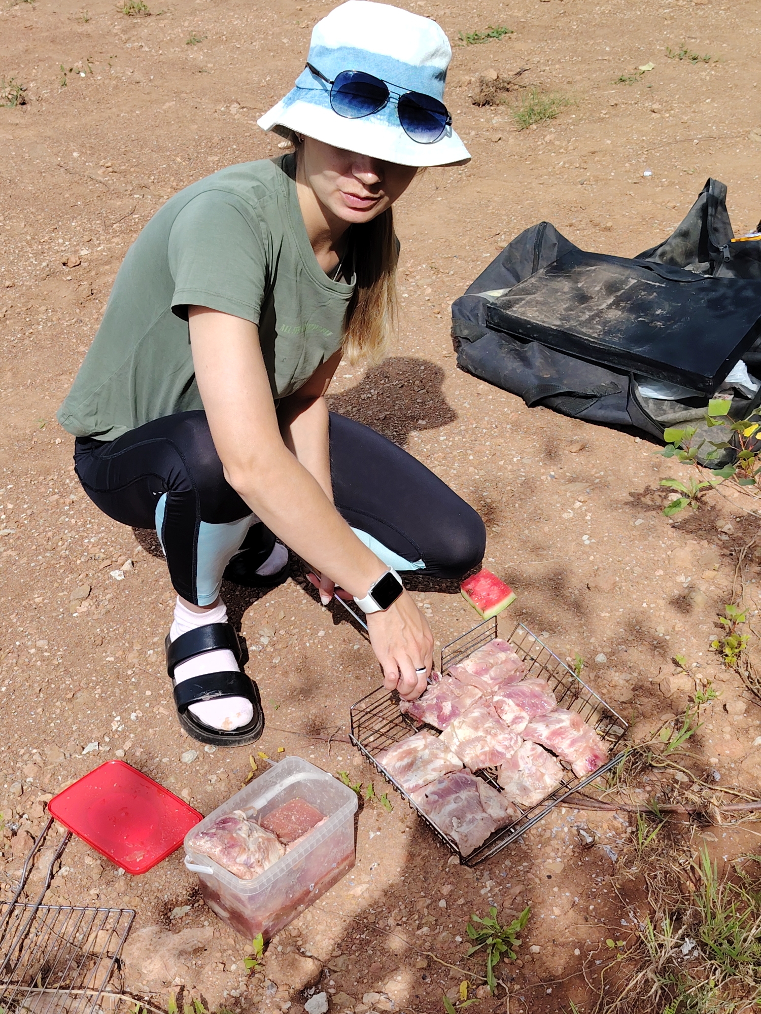 A young woman wearing a bucket hat and sunglasses kneels on the ground in an outdoor, likely rural or park setting, in Прикамский, Russia. She is in the process of preparing raw meat for grilling. She wears a green t-shirt, black leggings with light blue accents, and black sandals with white socks. On her left wrist, she wears a smartwatch. In front of her is a metal grill basket filled with raw meat pieces. To her left, there is a red plastic container with a lid and a clear plastic container holding more raw meat. To the right of the grill basket, a slice of watermelon rests on the dirt. Behind her, a large black bag is visible. The ground is covered in dirt and scattered rocks, with sparse vegetation. The lighting suggests it is daytime.