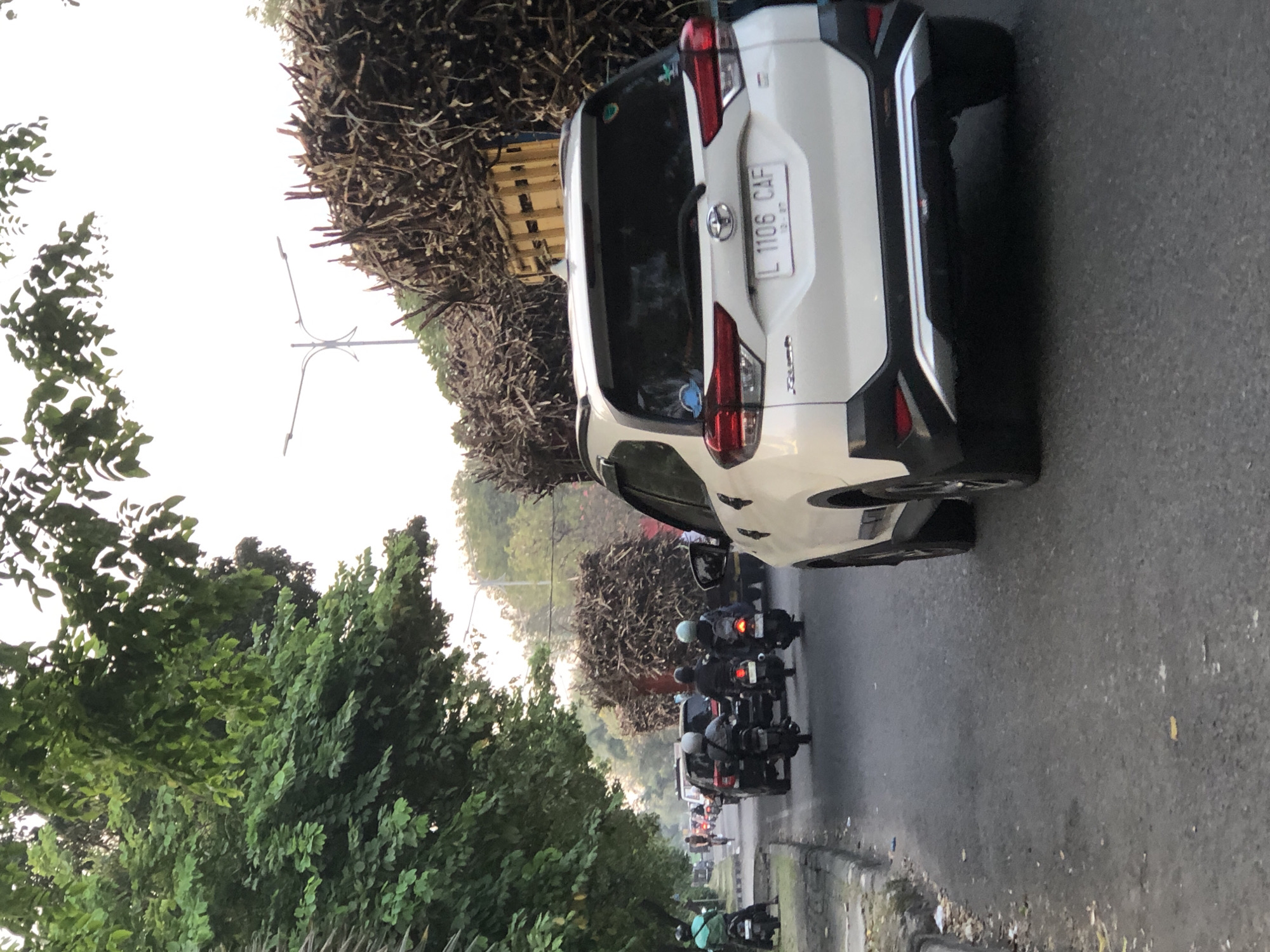 In Sumorame, Indonesia, on a paved road with a curb and sparse litter, a white SUV with license plate "L 1106 CAF" is positioned on the right side of the frame, facing away from the viewer. The SUV has a silver trim around its rear bumper and appears to be in traffic. To its left, a large truck, its bed filled with bundles of harvested sugarcane stalks, is also traveling in the same direction. The truck's yellow bed railing is visible. In the foreground, several motorcycles are present, three of which are in the middle of the lane behind the SUV. One motorcycle has a rider wearing a green shirt, another has a rider in a grey shirt, and the third has a rider in a light blue helmet. Further ahead, there are other vehicles, including a dark-colored SUV, and a person on a bicycle. Lush green trees with dense foliage line the left side of the road, with their branches extending over the street. The sky is overcast and bright, suggesting it is either early morning or late afternoon.