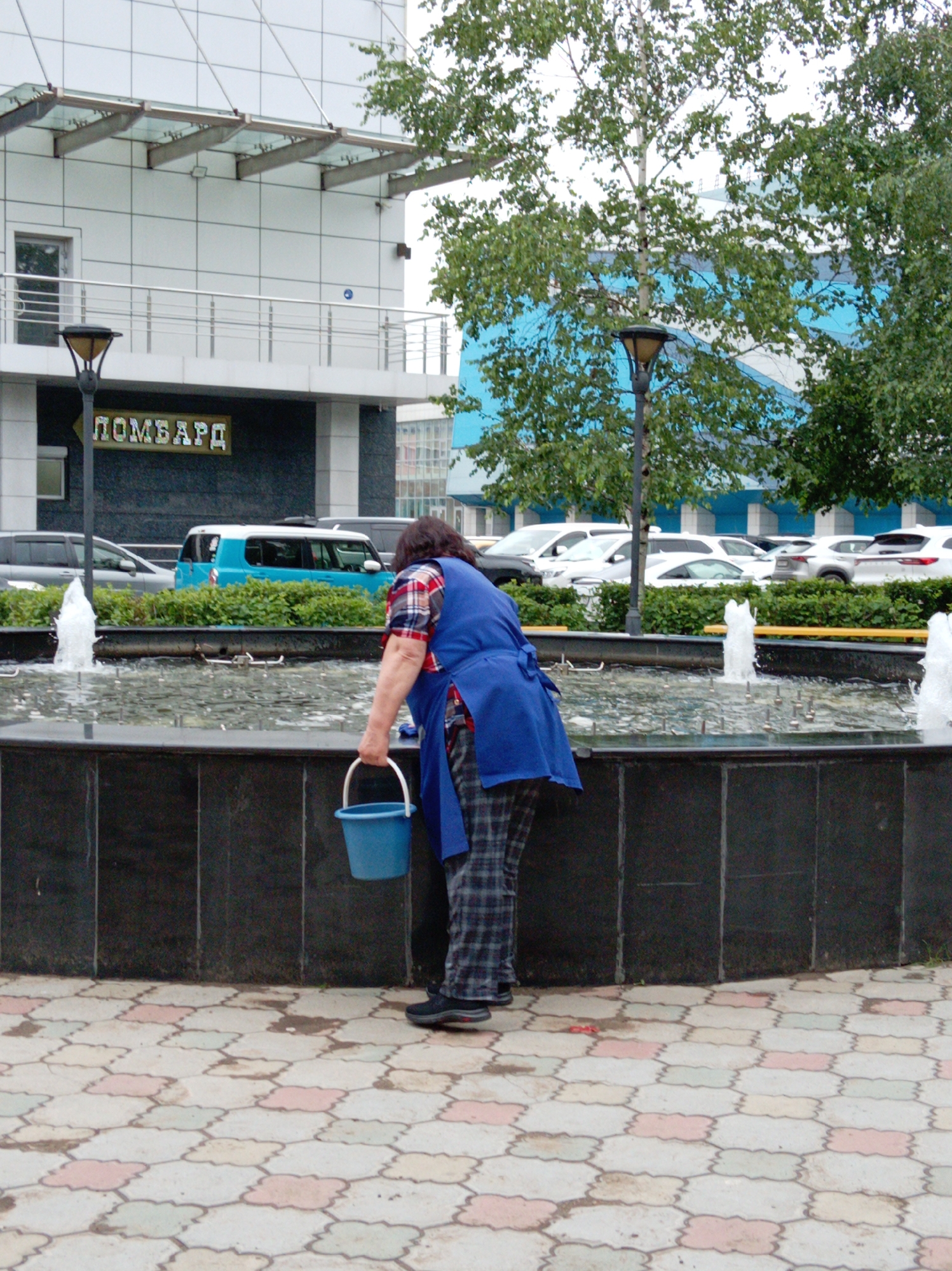 In Artyom, Russia, a person wearing a blue apron and plaid pants is cleaning a fountain. The person is holding a blue bucket in their right hand. The fountain has water jets and is surrounded by a dark granite base. In the background, a blue and white building is visible, with a sign that reads "Ломбард" (Lombard). Several cars are parked in front of the building. The ground is paved with interlocking tiles.