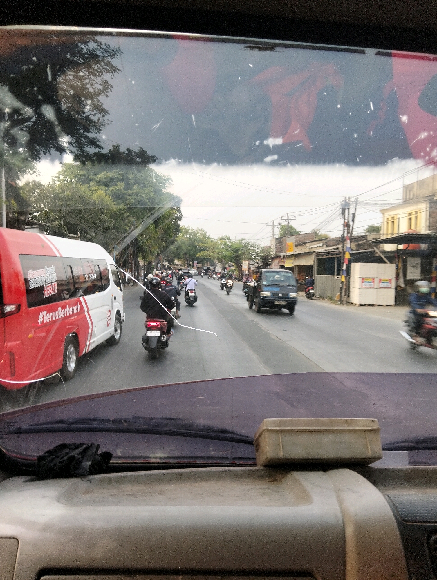 The image shows a view from inside a vehicle looking out onto a street in Semarang, Indonesia. A red and white bus with the text "#TerusBerbenah" is visible on the left side of the road. Multiple motorcycles and a small blue truck are also on the road, suggesting traffic. Buildings line the right side of the street, with power lines and trees interspersed. The sky is overcast. Inside the vehicle, a black glove rests on the dashboard in the foreground.