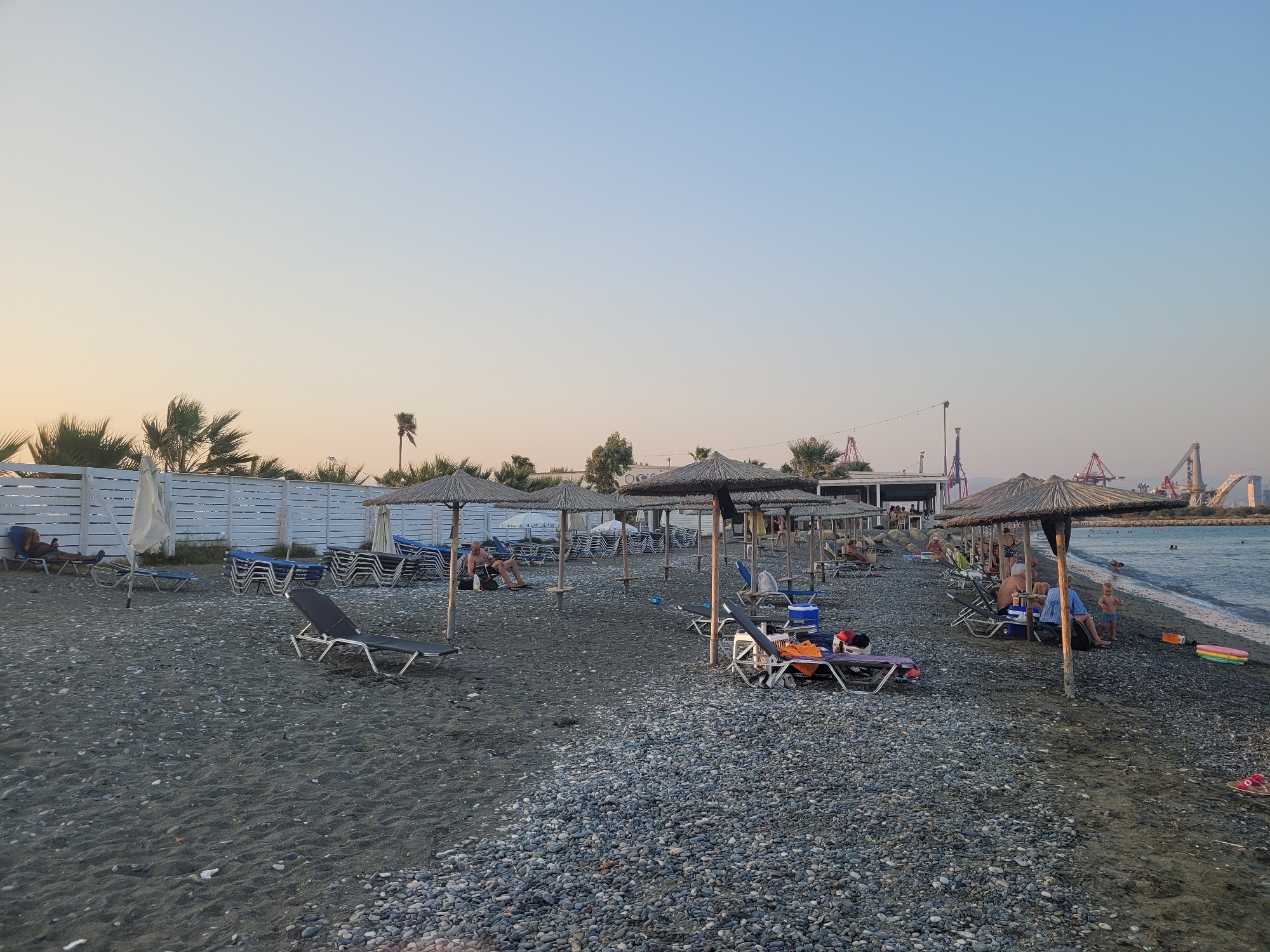 The image depicts a seaside scene on a pebble beach with patches of sand. Numerous beach chairs and straw umbrellas are arranged on the beach, some occupied by people. In the background, a white wooden fence runs along the edge of the beach, with palm trees and other vegetation visible behind it. The sea is calm, with a few people swimming or wading in the water. In the far distance, port cranes are visible, suggesting the presence of a harbor or industrial area nearby. The sky is a gradient of pale blue and orange, indicating either sunrise or sunset. The overall atmosphere is peaceful and relaxed, with the typical ambiance of a beach resort.