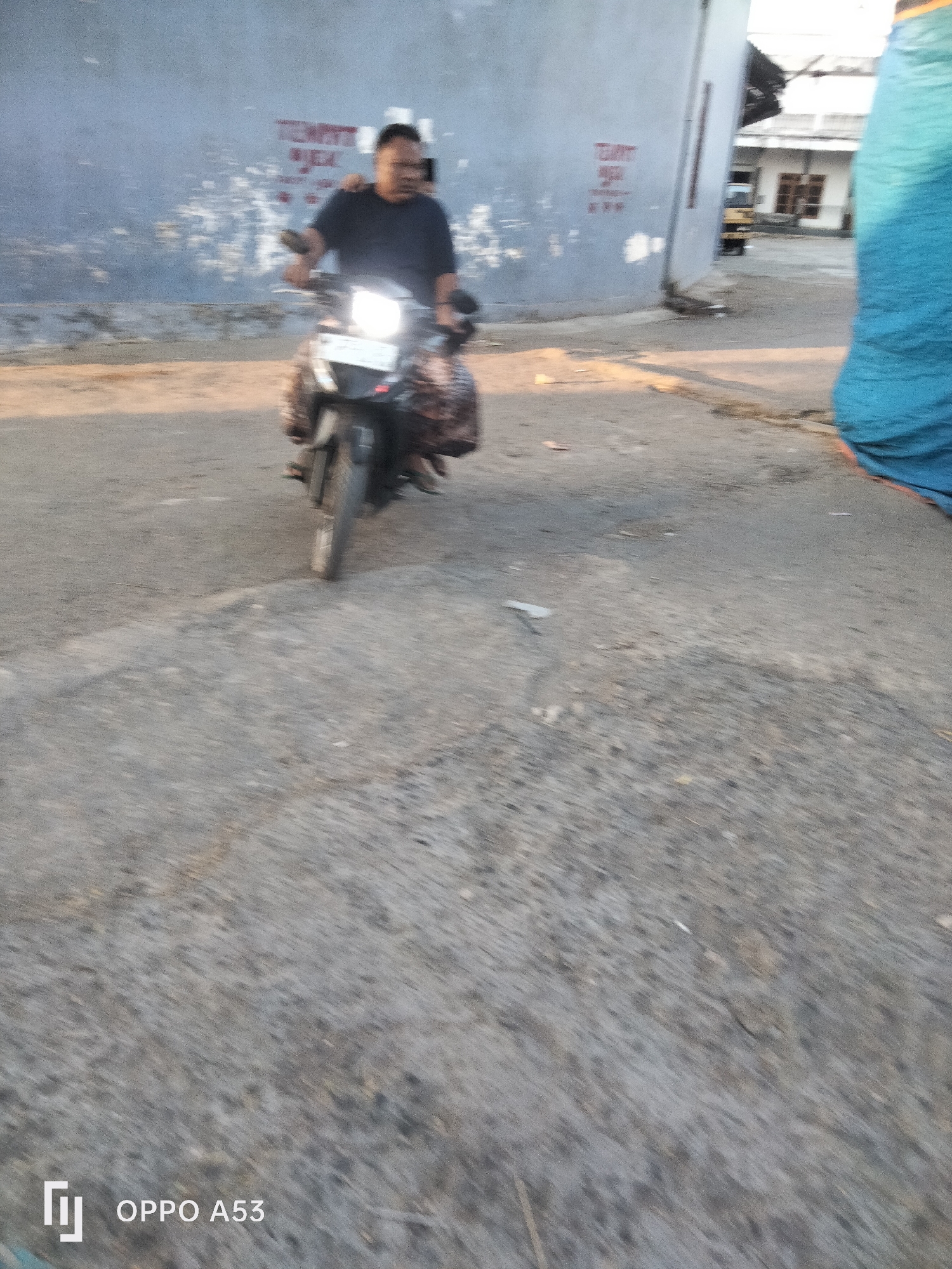 A man rides a motorcycle down a street in Badung, Indonesia. The man is wearing a dark t-shirt and is looking ahead. He is on a black motorcycle with a bright headlight. The street is paved with asphalt and is lined with a blue wall on the left and a building on the right. There is a large blue tarp draped over something on the far right of the image. The lighting suggests it is either early morning or late afternoon. There are some markings on the blue wall that are not clearly discernible. The ground has some debris scattered on it. The image has a watermark in the bottom left corner that reads "OPPO A53".