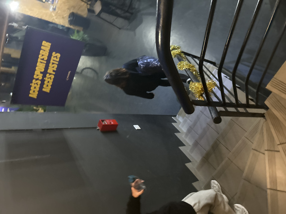 From an elevated perspective, a person walks down tiled stairs beside a black metal railing adorned with gold tinsel. A sign labeled "ACCÈS SPORTSBAR ACCÈS PISTES" stands to the left. Adjacent to it, a red box-shaped object and a small white card are positioned on the wall. Another person, holding a water bottle, is partially visible at the bottom of the frame.