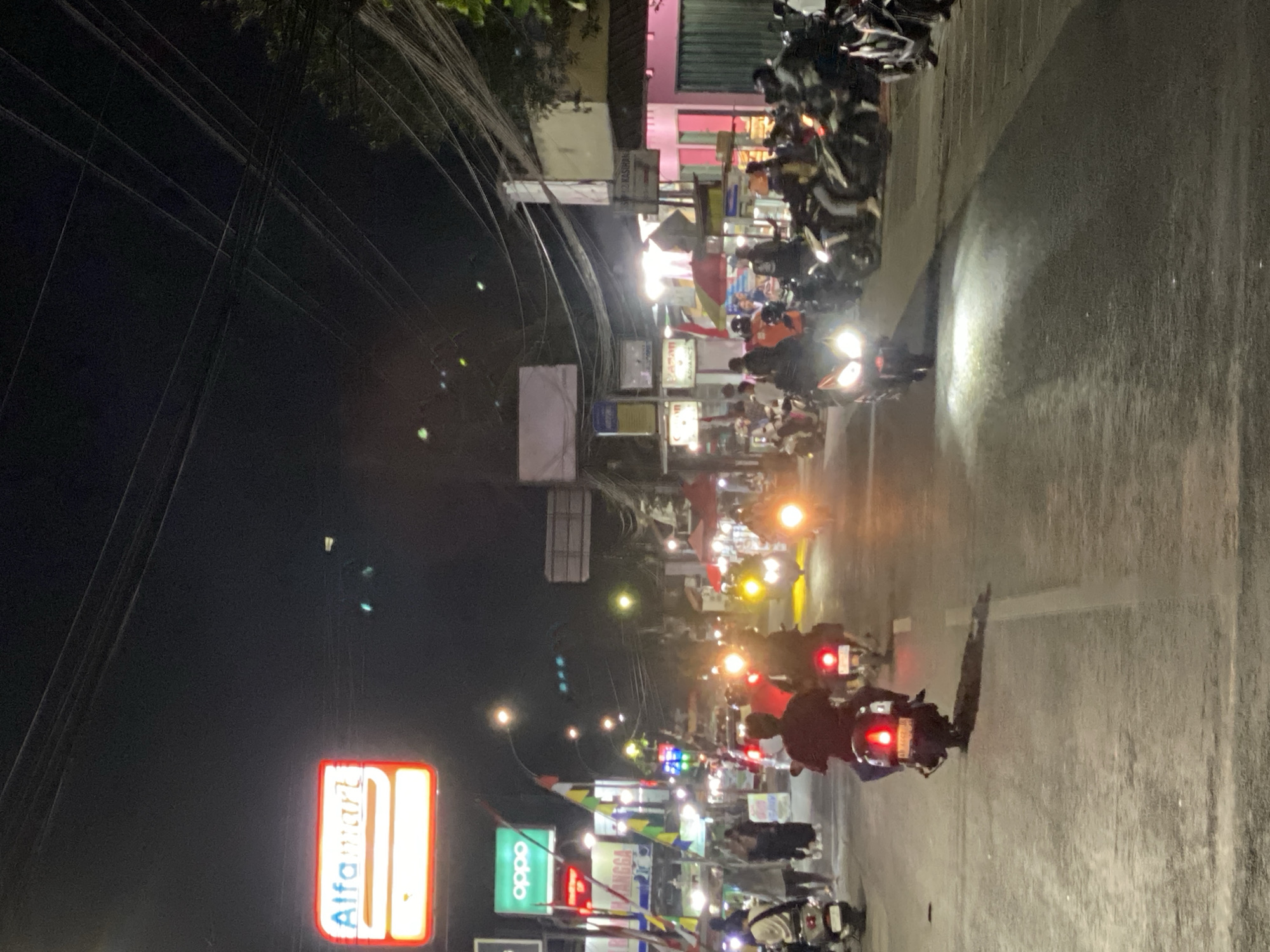 This image depicts a street scene at night in Kasihan, Indonesia. The road is asphalt and appears wet, with visible lane markings. Several motorcycles are present, some occupied and in motion, their headlights illuminating the road. On the left side of the street, there is a red and white illuminated sign for "Alfamart," a convenience store, and a green illuminated sign for "Oppo." Further down the street, there are various shops and stalls, some with awnings and lights, creating a busy commercial atmosphere. Pedestrians are visible walking on the sidewalks. The overall environment suggests a typical evening in a well-trafficked urban or suburban area, characterized by nighttime commerce and a significant amount of vehicular and pedestrian activity, primarily centered around the numerous motorcycles. Overhead, a complex network of electrical wires is prominent against the dark sky.