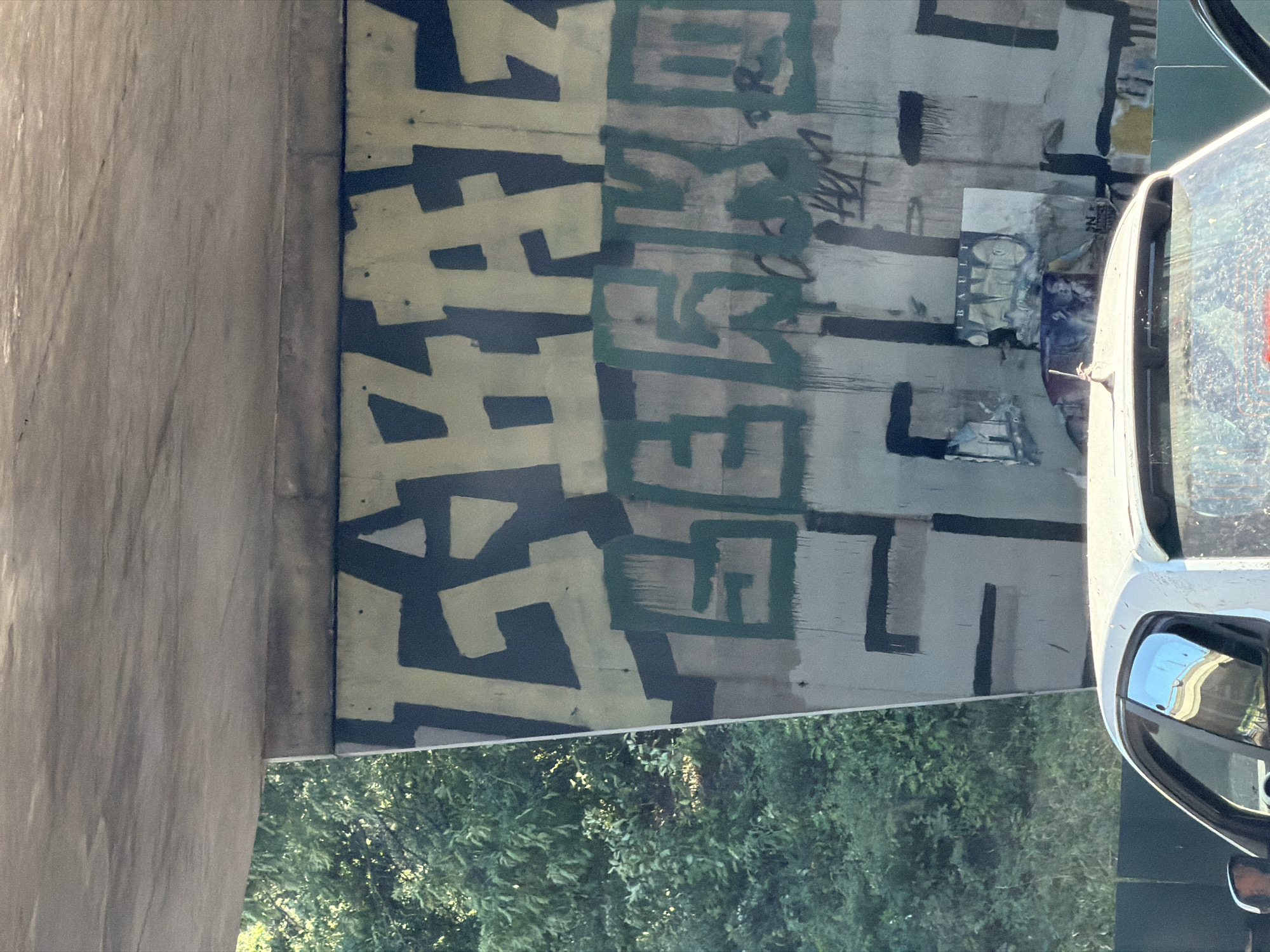 The image shows graffiti on a concrete structure, likely an overpass or bridge, with trees and foliage in the background. A white car is partially visible in the foreground. The graffiti consists of large, stylized lettering in light yellow and dark green on a grey and dark blue background. There are also some posters or paper remnants attached to the concrete wall below the graffiti. The overall scene appears to be an outdoor urban environment.
