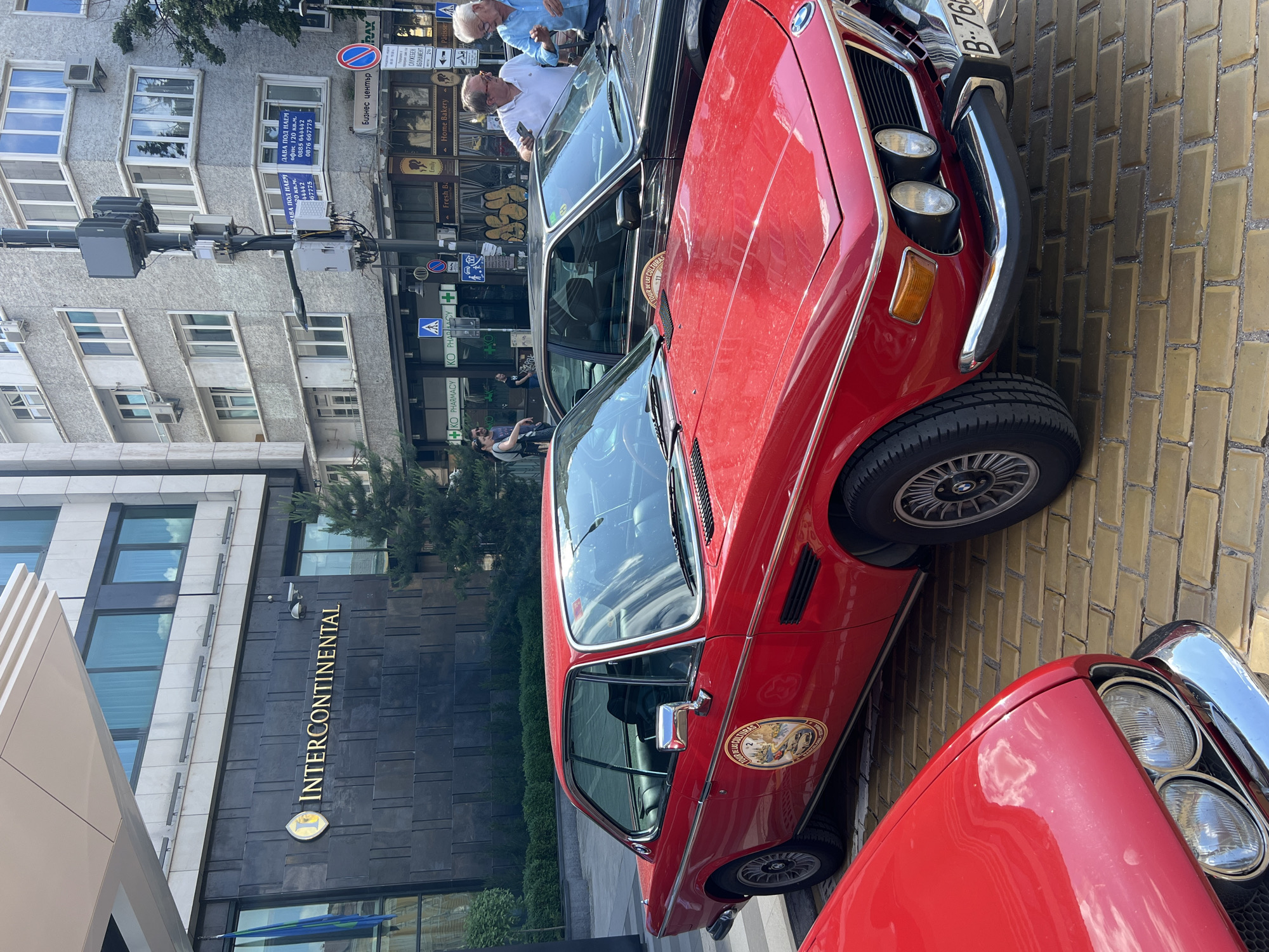 The image depicts an urban street scene in Sofia, Bulgaria, featuring two classic BMW cars parked on a paved surface. In the foreground, a glossy red BMW coupe, likely a 1970s model, is prominently displayed. It has chrome bumpers, dual headlights, and a sticker on its side door reading "TOUR DE LAS CULTURAS" with a landscape graphic. Part of its Bulgarian license plate, "B-760", is visible, where 'B' denotes Sofia. Behind it, a dark-colored classic BMW coupe is also parked, displaying a similar sticker.
Several individuals are present, mostly adult males. Two older men are conversing near the dark BMW, with one holding a smartphone. Another man in a white polo shirt is visible behind them, and further back, a woman and another man are walking.
The background features several buildings. To the left, the dark facade of the Intercontinental Hotel is visible, with its golden "INTERCONTINENTAL" sign and logo clearly displayed. A blue and green flag hangs near the hotel entrance. To the right of the hotel, an older, light grey multi-story building with numerous windows and some external air conditioning units dominates the mid-ground. On the windows of this building, several blue "ДАВА ПОД НАЕМ" (FOR RENT) signs are posted in Cyrillic, along with phone numbers and "офис 120 кв.м." (office 120 sq.m.). At street level, various retail establishments are present, including "KO PHARMACY," "Emily," "Home Bakery," and "Patisserie." A street sign in Cyrillic for "Бизнес център" (Business Center) is also visible. A tall, multi-component utility pole stands in the center-right of the frame. The street is paved with light brown or yellowish rectangular bricks. The scene is illuminated by daylight under what appears to be a clear sky, with reflections of clouds visible on the car surfaces and building windows.