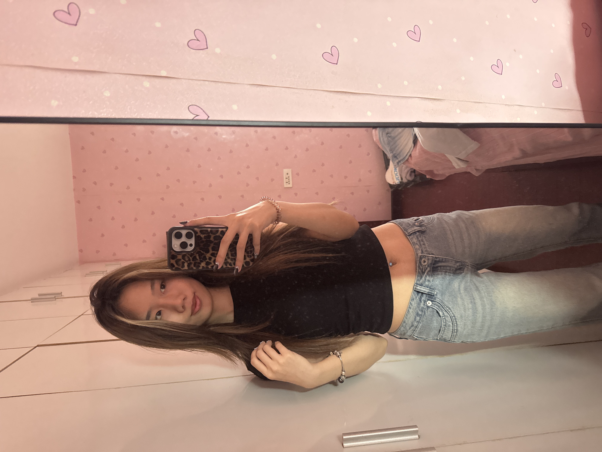 A young woman poses for a selfie in a mirror. She is wearing a black top and light blue jeans. She has long brown hair with blonde highlights. She is holding a phone with a leopard print case. The background shows a pink room with heart-patterned wallpaper. The location appears to be a residential setting, possibly a home, within Shapingba District, China.