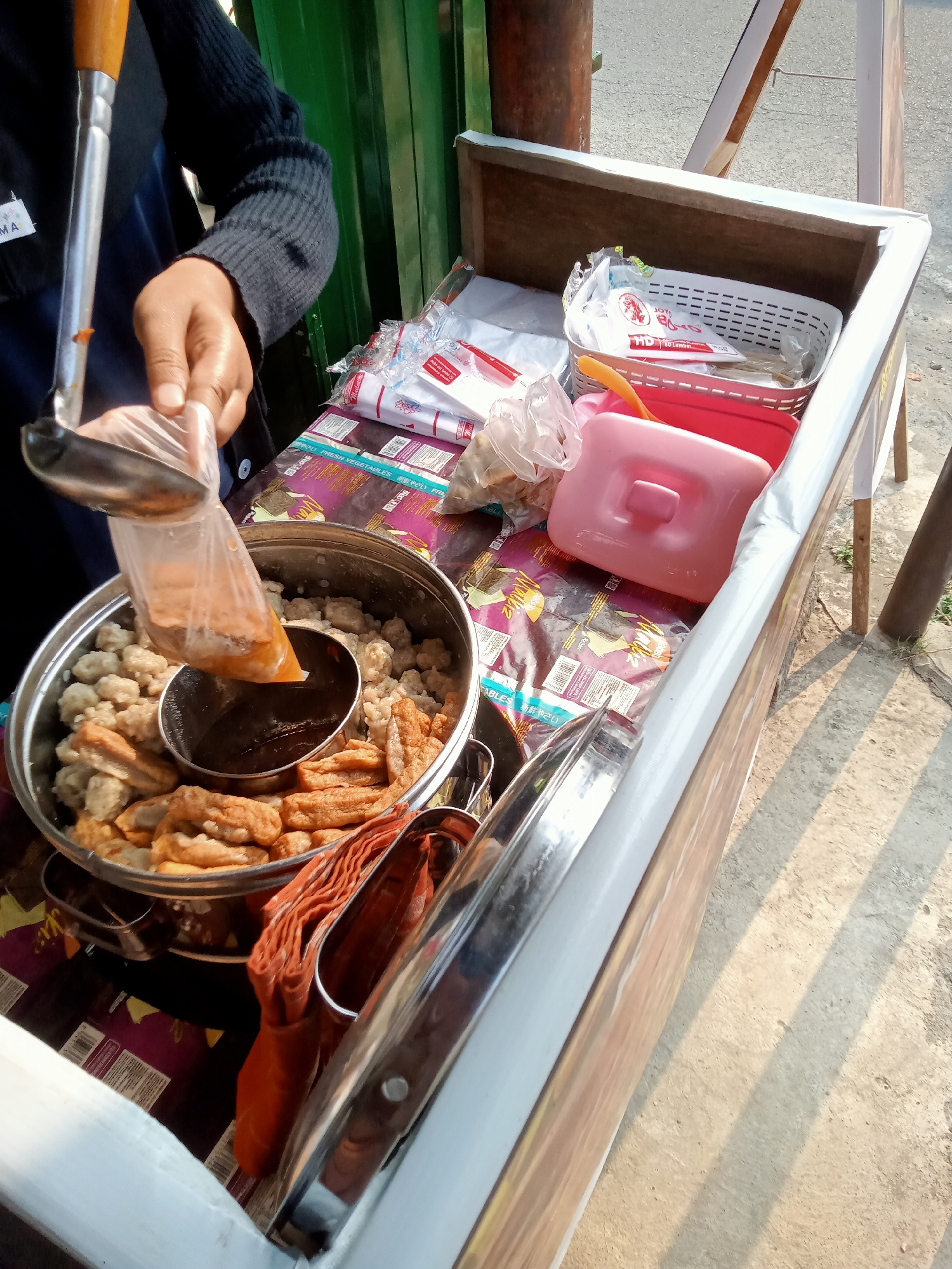 A food vendor in Simo, Indonesia, is seen preparing a serving of street food from a cart. The cart is laden with various food items and serving utensils. In the foreground, a large stainless steel pot is filled with round, light-colored meatballs and rectangular, fried pieces of what appears to be tofu or fish cake. A smaller metal bowl sits within the larger pot, containing a dark, viscous sauce. The vendor's gloved hand is holding a clear plastic bag filled with the sauce, which is being poured into the central bowl using a metal ladle with a wooden handle. Behind the pot, various food packaging and plastic containers are visible. The cart itself appears to be constructed from wood with a white lining. The scene is set outdoors, with a concrete surface visible in the lower portion of the image, suggesting a street or market setting. The lighting indicates it is daytime, with shadows cast across the ground.