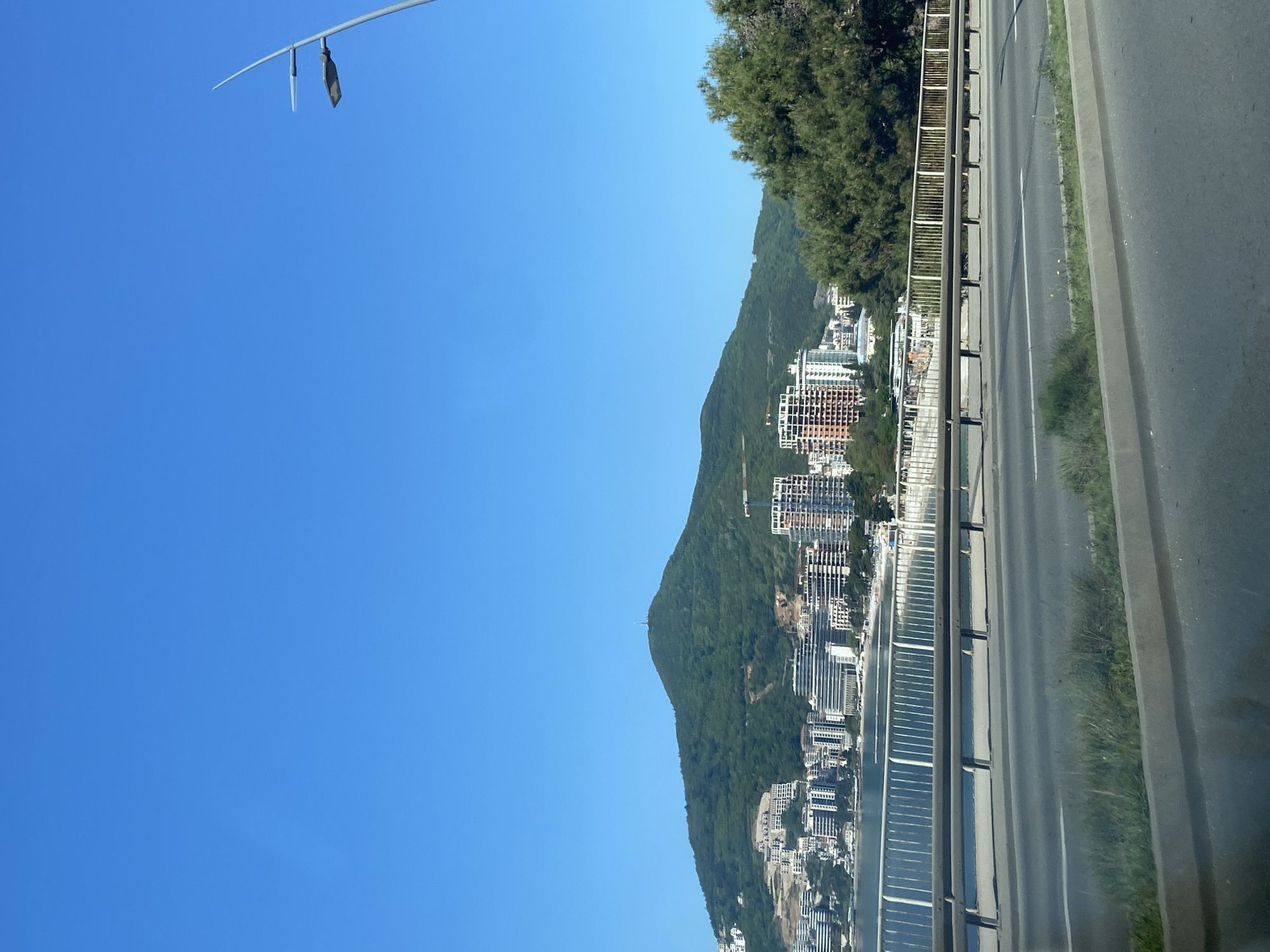 The image captures a scene in Budva, Montenegro, showing a coastal road running alongside a hillside urban development on a sunny day. The sky is clear blue. A portion of a lamppost is visible at the top left corner of the frame. To the right, the hillside rises with a mix of green vegetation and numerous high-rise residential buildings. The buildings appear to be a mix of modern architectural styles, with multiple floors and balconies. The road is in the foreground with a metal railing separating it from the hillside. Grass and concrete road features can be seen on the road.