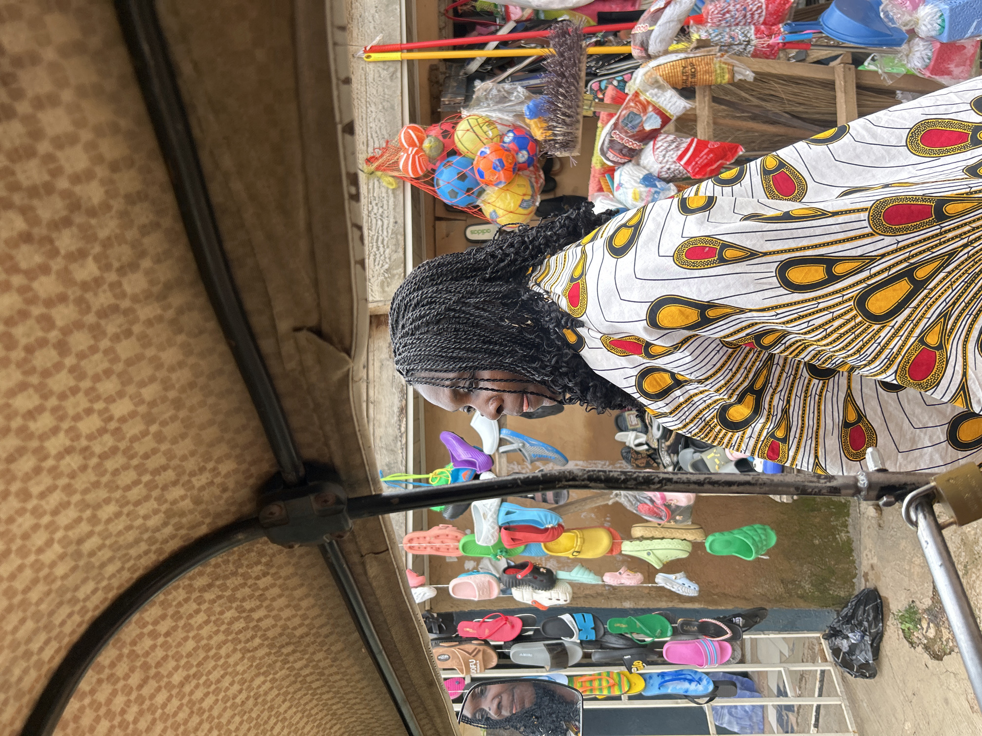 The image shows a woman with braided hair wearing a white dress with a colorful peacock-feather-like pattern. She is standing in what appears to be an outdoor market stall in Jos, Nigeria. In the foreground, a canopy made of tan fabric with a checkered pattern is visible, supported by a dark metal frame.
The woman is looking downwards, and her reflection is visible in a mirror placed on a metal railing in front of her. Behind her, the stall is filled with various items. To her left, a net bag holds several colorful balls, some of which are blue, orange, and yellow. Further back, there are cleaning supplies such as brooms and mops, many still in their packaging.
The stall also displays a wide assortment of footwear, including sandals, flip-flops, and clogs in various colors like pink, yellow, green, blue, and red. These are arranged on shelves. Some of the shoes have visible brand names or text, such as "TOFU" and "adidas". The overall impression is of a busy market scene, possibly during daylight hours. The ground appears to be a dirt or concrete surface, and there is some water or dampness visible.