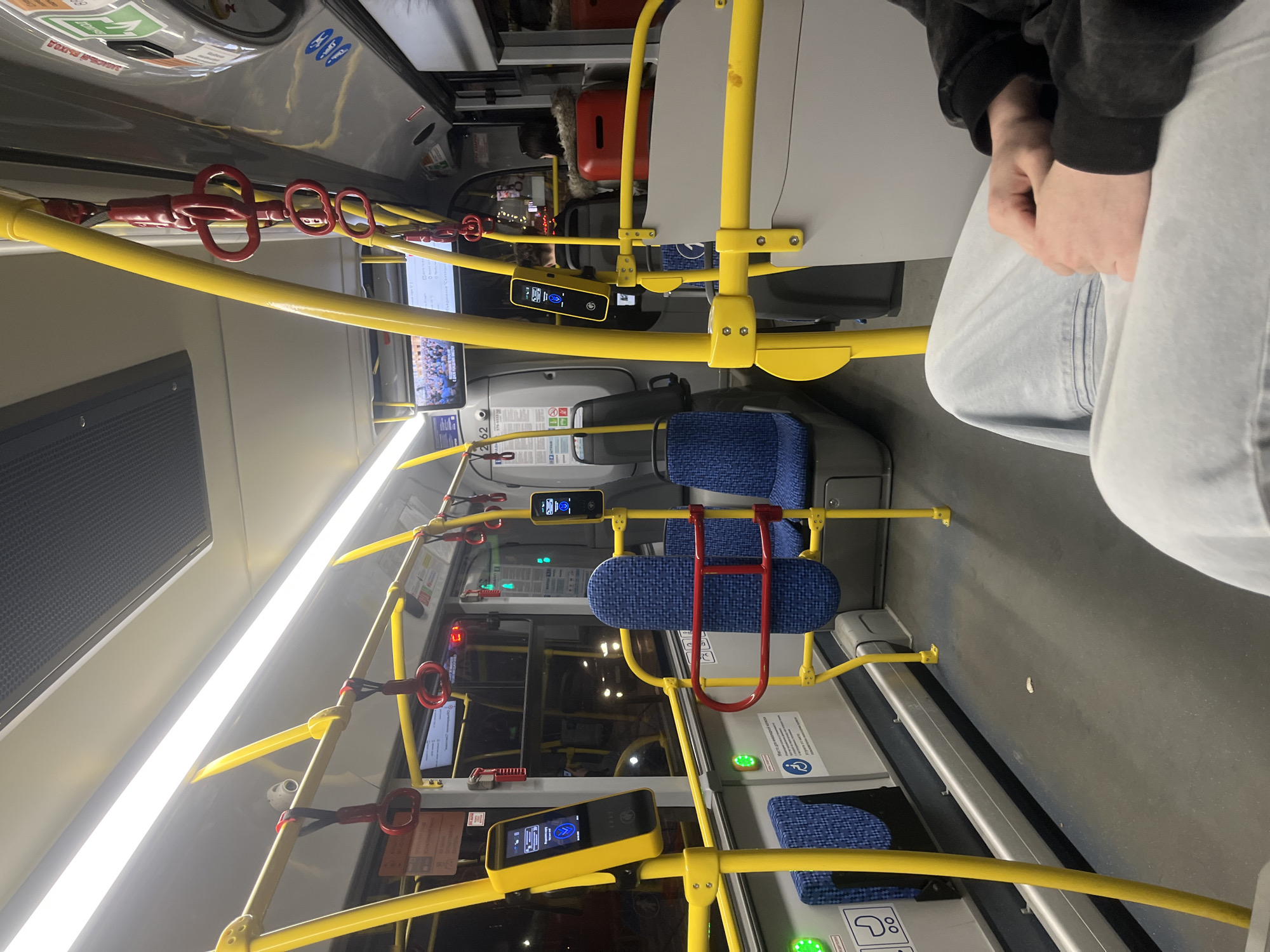 The image depicts the interior of a public bus, likely in Saint Petersburg, Russia, as evidenced by the Cyrillic script on signage and display screens. The seating arrangement is characterized by blue, patterned fabric. Yellow handrails equipped with red hand-holds are visible throughout the bus. A person is seated in the right foreground, wearing light-colored jeans and a dark jacket. Various electronic information displays are attached to the handrails, likely for passenger information. The lighting is bright and appears to be mostly artificial. The bus is in motion, suggested by the blurred background.