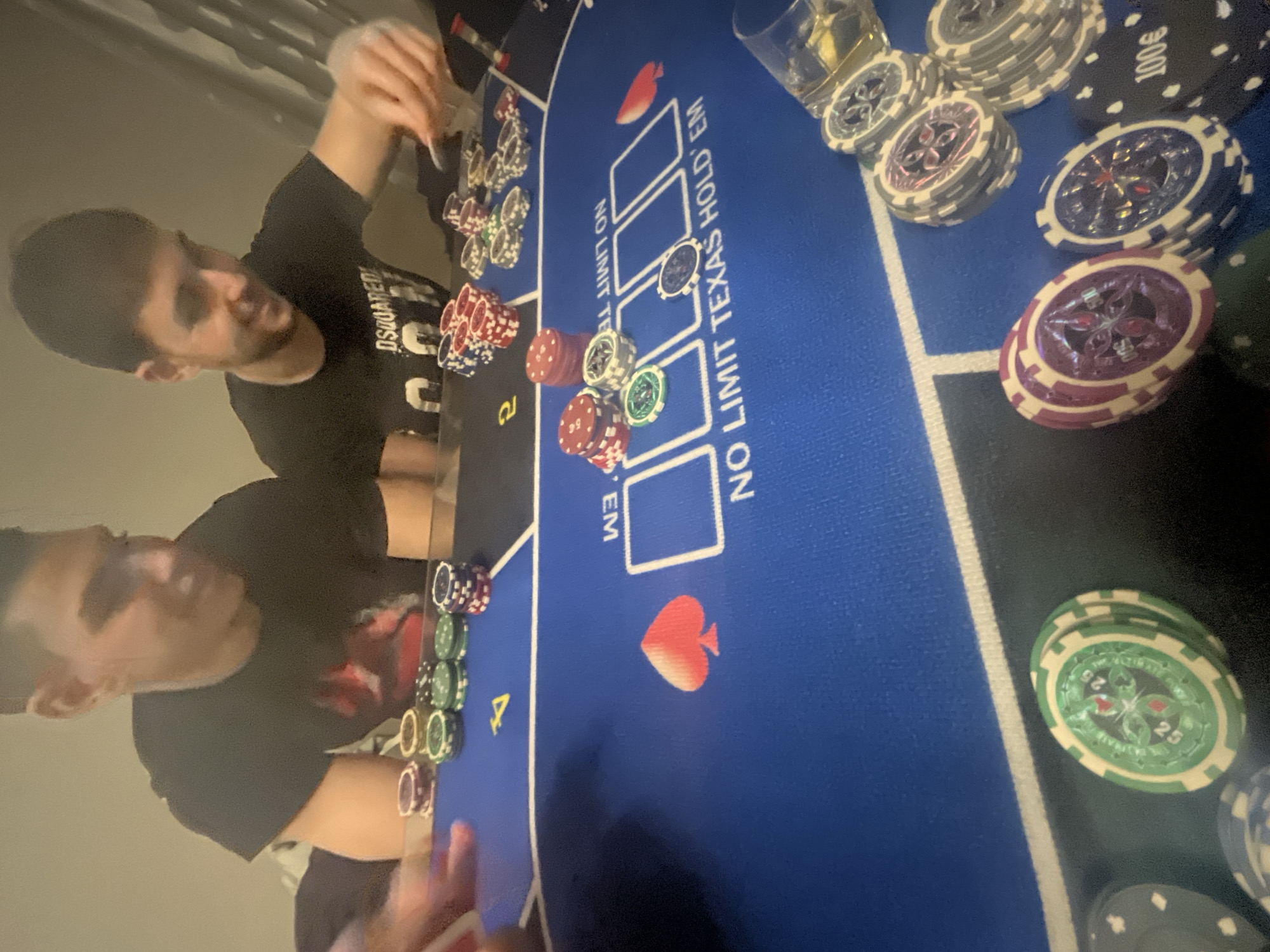 Two men are at a blue poker table with the words "NO LIMIT TEXAS HOLD'EM" printed on it, with poker chips and a glass with a yellow liquid on the table. One man is gathering chips with his right hand, and the other man is looking at the chips. The chips are various colors. The table also has card suit symbols printed on it. The image does not contain any information that indicates it was taken in the city of Burgas, Bulgaria.