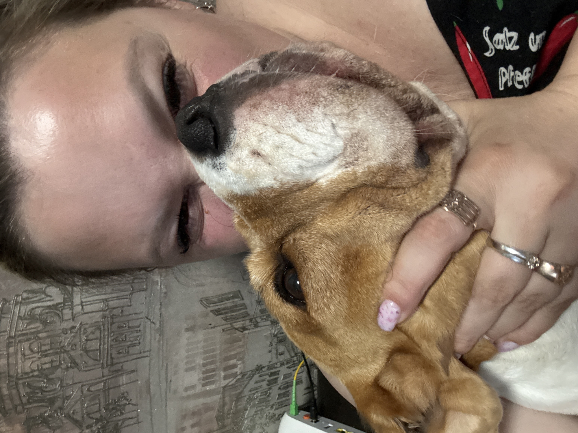 A woman is lying down indoors, embracing a tan and white beagle. The woman has closed eyes and dark eyelashes. Her right hand is holding the dog's head. The woman wears rings on her fingers. A black garment with white text ("Satz un Pfeff") is visible on the woman's upper right. Below the dog's head, a white electronic device with green and yellow cables is visible. The background surface appears to be patterned. The location is Марковский, Russia.