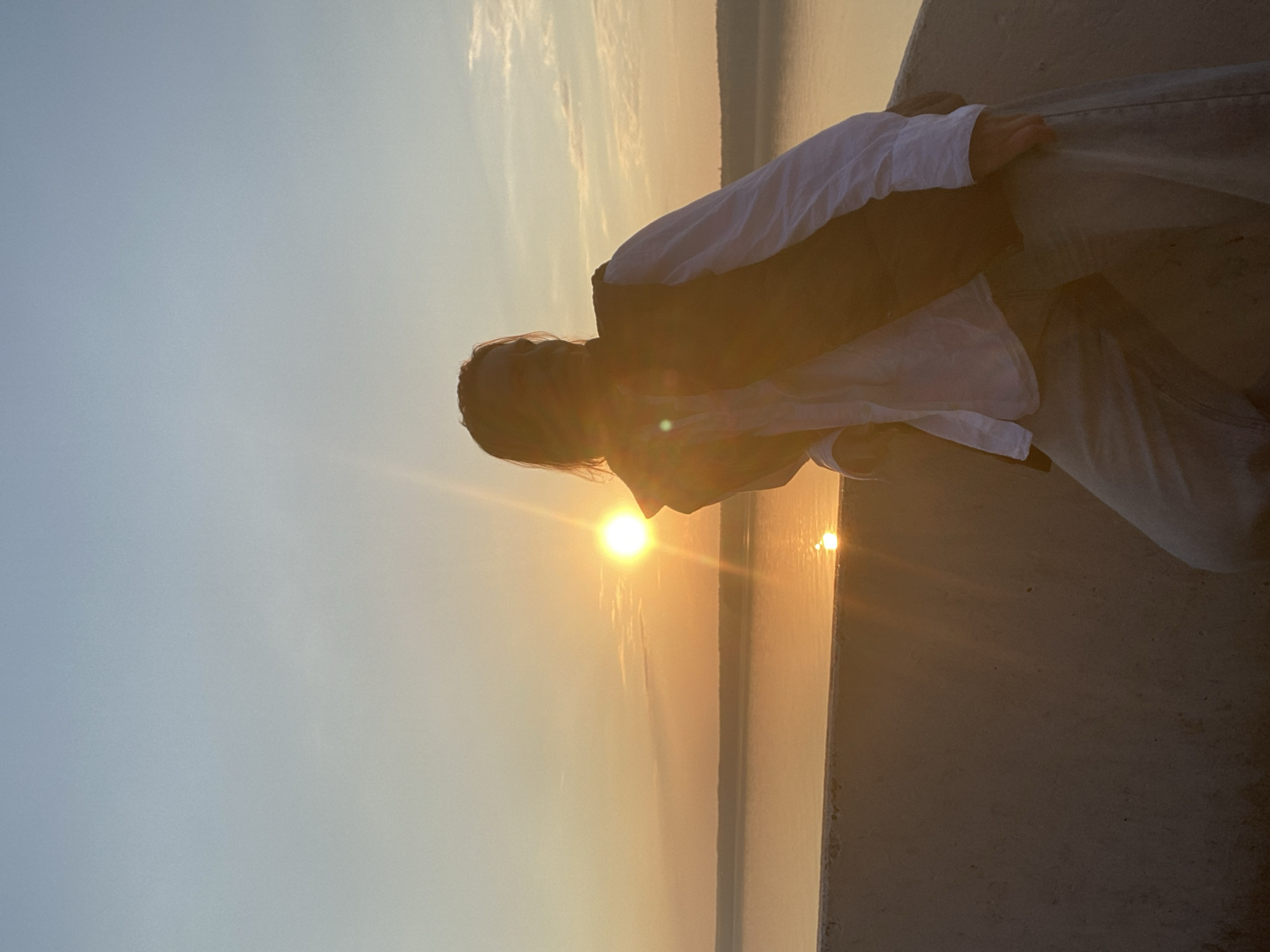 A person is standing outside, likely at sunset, in Chaikovsky, Russia. The person is wearing light-colored pants, a white collared shirt, and a dark vest. The sun is low in the sky, creating a bright glare and a reflection on the water. There is a large concrete structure or wall to the right of the person.
