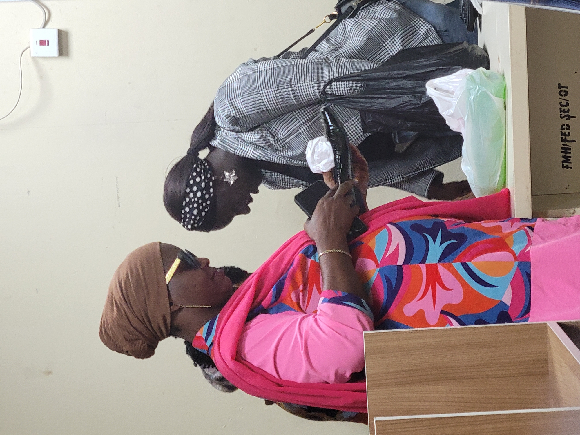 Two women are present. One is wearing a brown hijab, sunglasses, and a pink top with a colorful scarf. The second woman wears a black and white patterned jacket, a black head scarf with white dots, and earrings shaped like flowers. The woman in the hijab holds a bottle, offering it to the other. The background includes a light beige wall and what seems to be office furniture with text "FMH/FED SEC/OT". The location is Abuja, Nigeria.