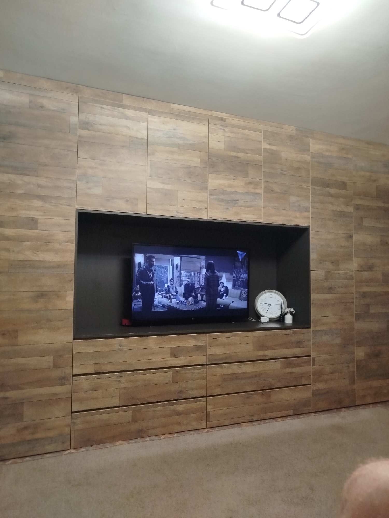 The image shows a living room with a large entertainment unit. The unit has a wood-grain finish and features a recessed area for a television. The television is on and displaying a scene with several people. To the right of the television, there is a white analog clock and a small white spray bottle. Below the television, there are several drawers integrated into the unit. The room is illuminated by a modern ceiling light fixture. The floor is carpeted. The scene does not provide any specific geographical markers to pinpoint a location within Naberezhnye Chelny, Russia, or any other city.