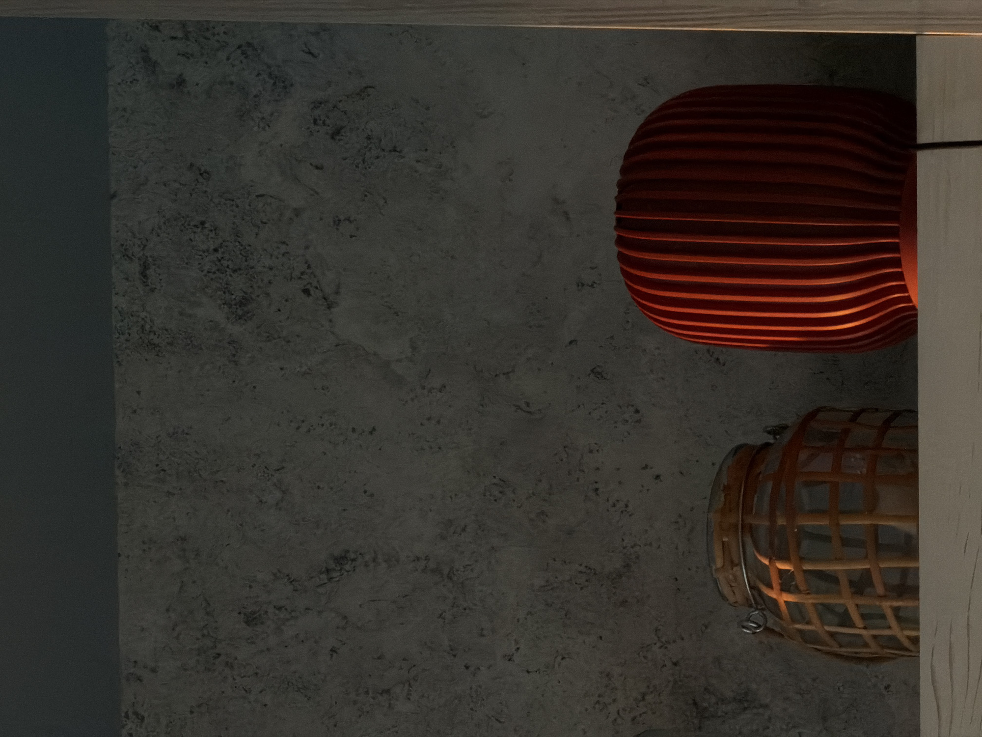 The image depicts a dimly lit interior with two decorative objects: a horizontally striped, reddish-orange lamp and a wicker basket-like object, both positioned against a textured gray wall. A dark gray surface is visible on the left side of the image, possibly part of a wall or panel. A light-colored wooden frame is on the right. There is no indication of people, specific actions, interactions, or the location in Khimki, Russia.