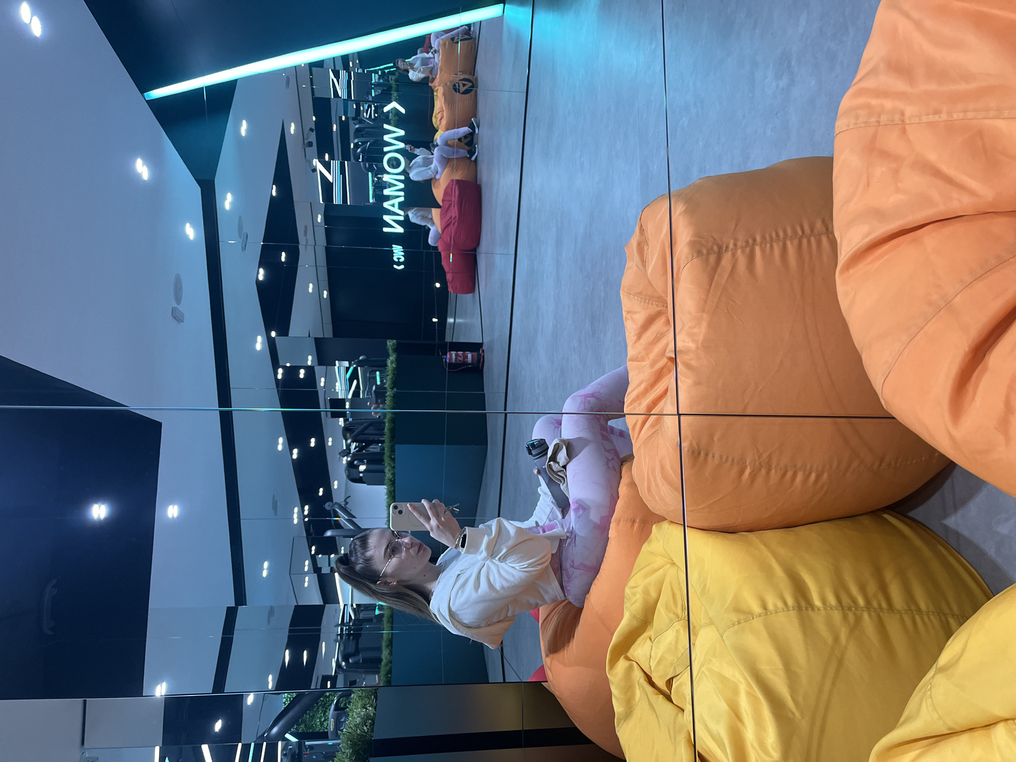 A woman with a ponytail, glasses, and white hoodie is sitting on a stack of large, colorful beanbag chairs (orange, yellow) and holding a smartphone, taking a picture of herself in front of a floor-to-ceiling mirror. The reflection shows a gym-like setting with exercise equipment, and a line of people sitting on similar beanbag chairs in the background. A sign reading "WOMAN" is visible in reverse on the mirror. The environment appears to be a modern, brightly lit space. The location is Unknown, Unknown.