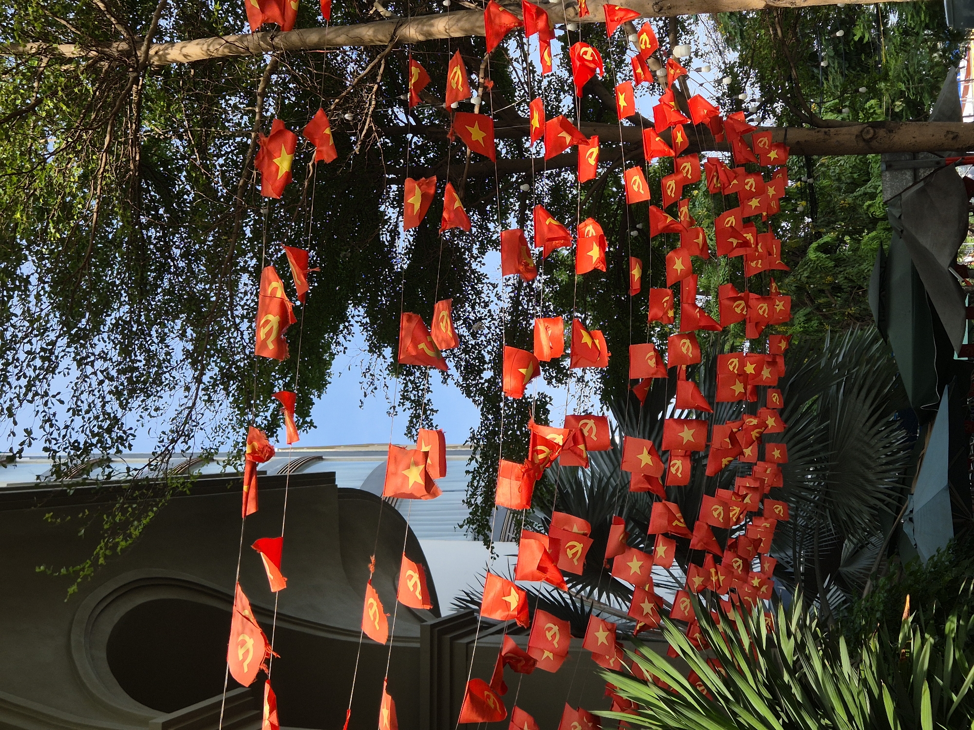 The image depicts a street in Đà Nẵng, Vietnam, adorned with numerous small red flags bearing a yellow star and a yellow hammer and sickle symbol, indicative of Vietnamese national and communist party flags. These flags are strung along wires, creating a festive or celebratory atmosphere. They are suspended between a modern building with large arched windows and trees with dense green foliage. In the foreground, lush green palm fronds are visible. The sky is a clear blue, with some sunlight filtering through the trees. Several large umbrellas, likely for outdoor seating, are partially visible at the bottom of the frame. The scene appears to be outdoors, during daylight hours, and suggests a public space or commercial area in the city. There are no people visible in the image.