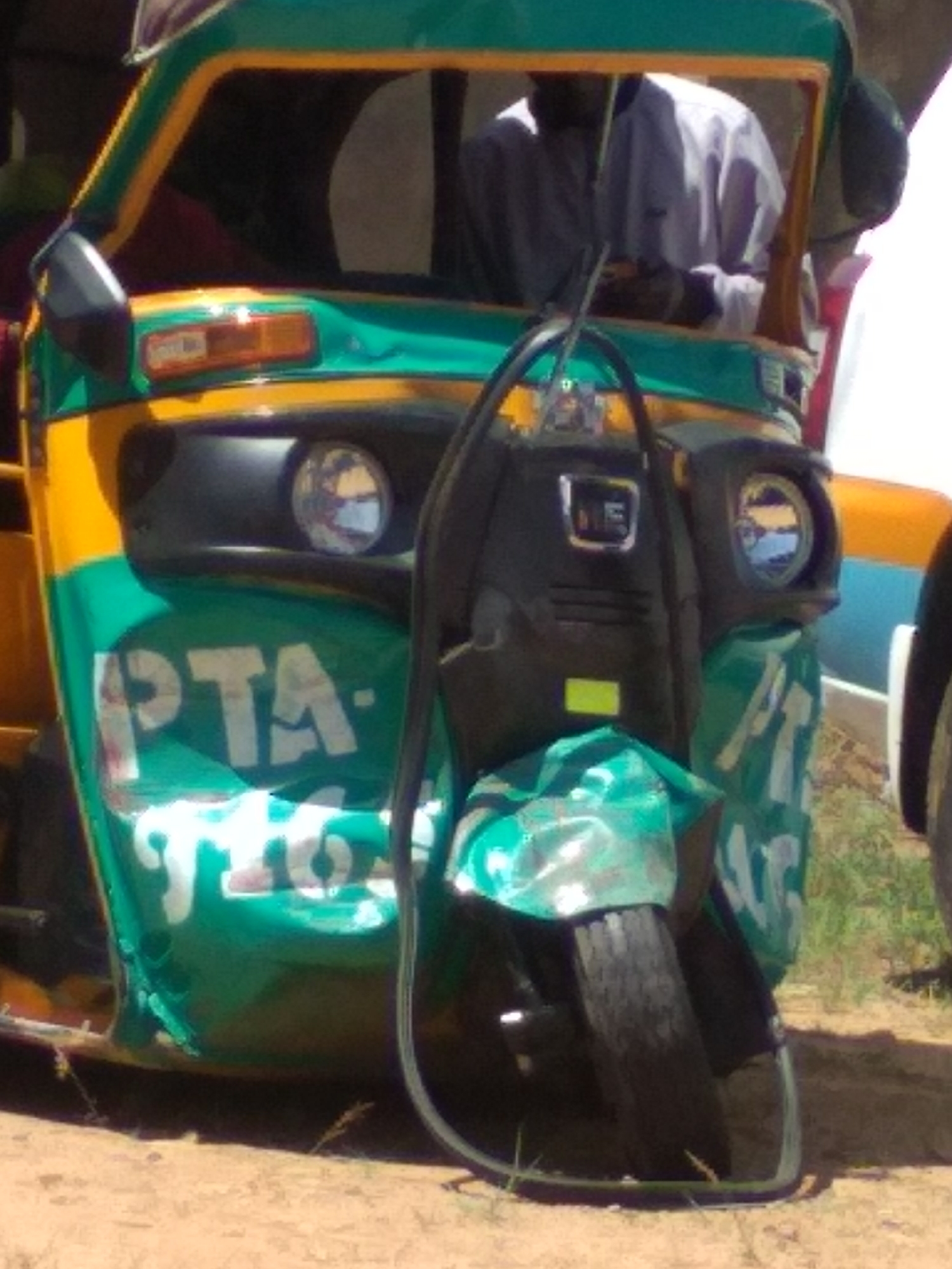 A partially obscured image shows the front of a green and yellow auto rickshaw, or tuk-tuk, parked on a dirt surface. The vehicle features a dark tire guard and headlights visible on either side of the front panel, which is painted with large, white letters that appear to spell out "PTA". A person wearing a light-colored shirt sits inside the rickshaw. The surrounding environment includes dry dirt ground and some sparse vegetation. The location appears to be a city in an unknown place.