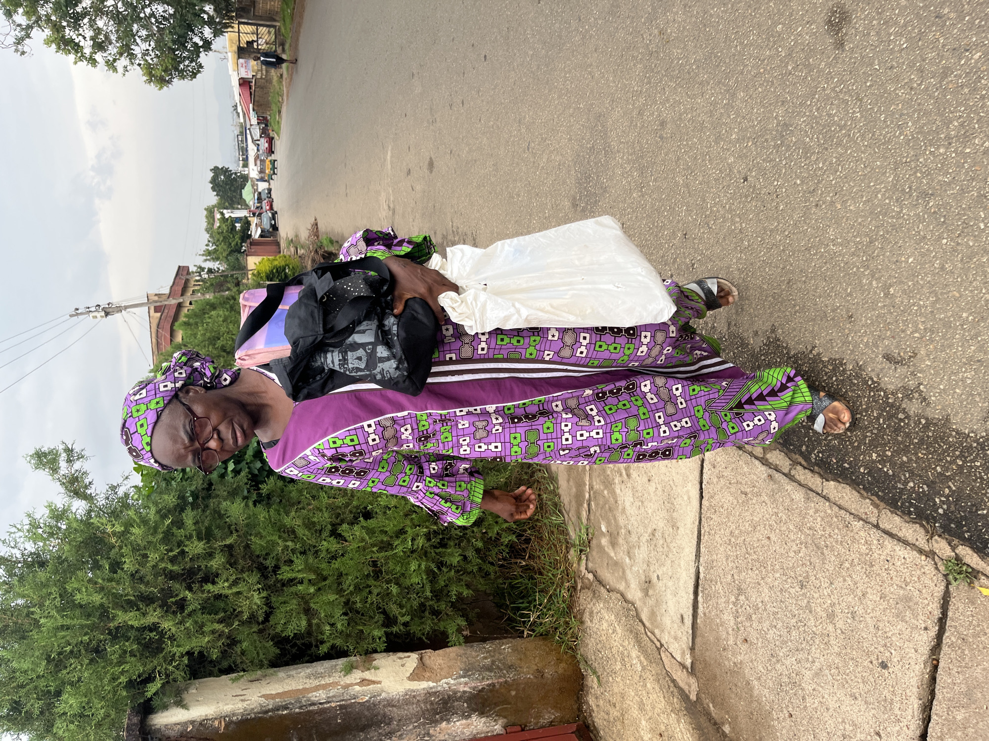 An individual wearing glasses, a purple and green patterned garment, and sandals is standing on a sidewalk. They are holding a white plastic bag and a black backpack. The location appears to be a city street, with vegetation present and vehicles visible in the background. The city and country are unknown.