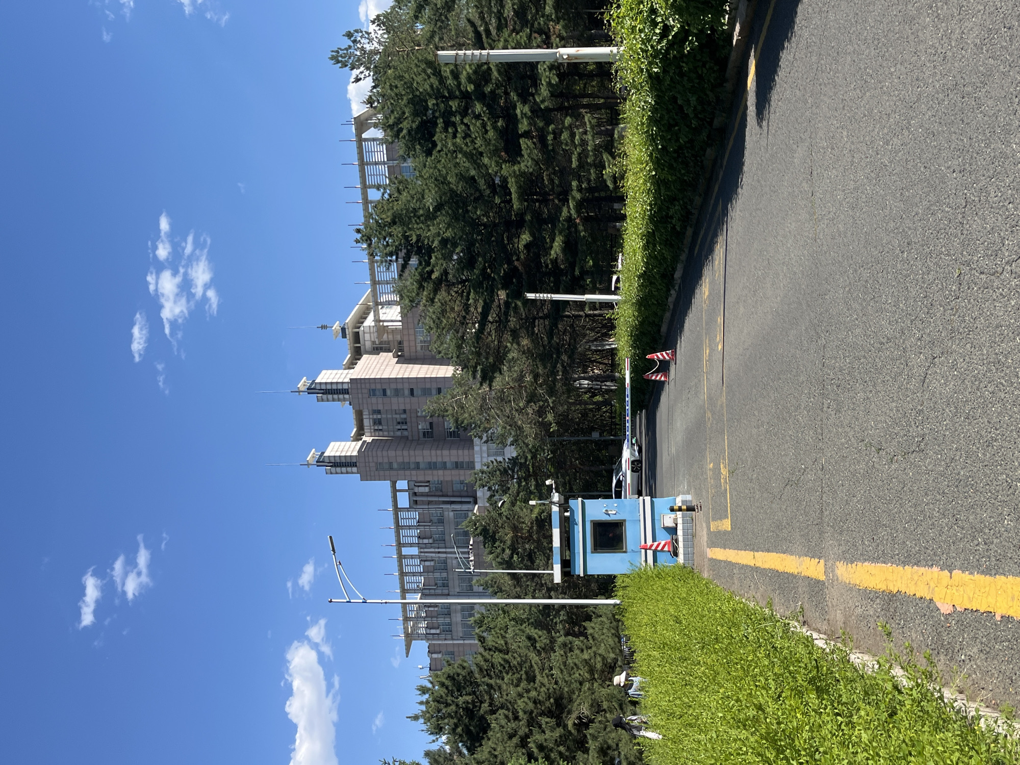The image shows a street in Changchun City, China, with a guard booth on the right side of the road. The booth is blue with white and red striped bollards next to it. Behind the booth, there is a barrier across the road. Tall buildings are visible in the background, obscured partially by trees. A streetlight is to the left of the guard booth. The sky is blue with scattered white clouds. The road is asphalt, marked with yellow lines. Hedges border the road. A car is faintly visible in the background, behind the barrier.