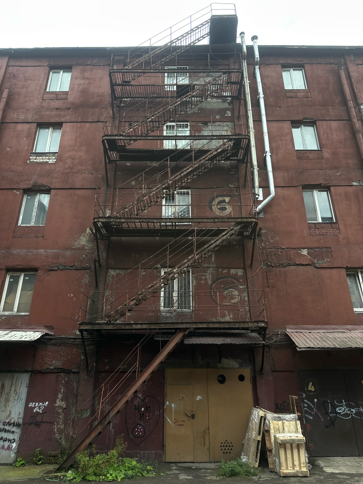 An exterior, low-angle, full shot shows a weathered, dark red, multi-story industrial building in Saint Petersburg, Russia. A rusty, black metal fire escape zigzags down the center of the building, with several landings and stairs. The building has multiple windows, some with white frames and others with dark frames, and some are barred. Two large metal pipes run vertically along the right side of the fire escape. The lower part of the building features several large garage doors. One garage door is yellow and has two circular openings, resembling eyes, and a pattern of small triangles below. Another garage door on the right is dark and has a yellow number '4' painted on it. The ground in front of the building is asphalt, with some weeds and green foliage growing. There is a stack of wooden pallets wrapped in plastic to the right of the yellow garage door. Graffiti is visible on some of the garage doors and on the lower part of the building's wall. The sky above is overcast and grey.