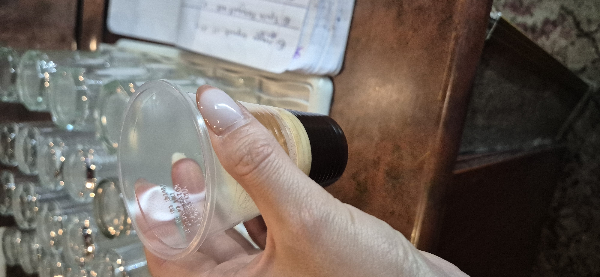 A person is holding a small glass with a black and gold colored lid, which appears to contain a white fluid, in their right hand. Numerous empty small glasses are arrayed to the left of the hand. The photo appears to have been taken on a wooden surface, with open notebook and a possible wooden dresser visible in the background. There is no information in the image to indicate a location in the city of Sofia, Bulgaria.