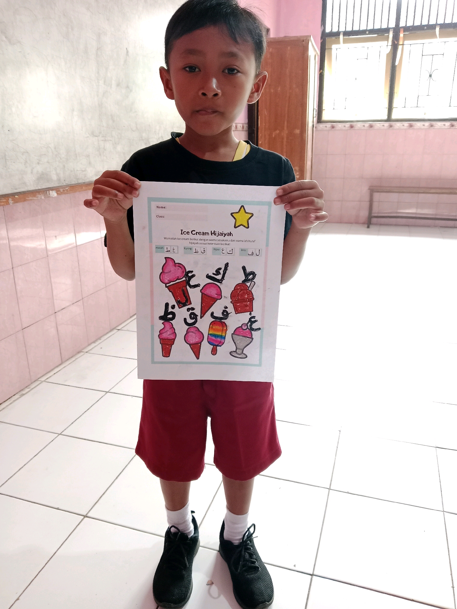 A young boy, likely of Indonesian ethnicity with short black hair and a neutral expression, stands in a well-lit room, holding up a worksheet titled "Ice Cream Hijaiyah." The worksheet features cartoon drawings of ice cream cones, each adorned with Arabic letters. The boy is wearing a black t-shirt, red shorts, white socks, and black athletic shoes. The room appears to be a classroom or similar educational setting, with tiled floors and a window with bars in the background. The walls are painted pink and white, and a wooden cabinet is visible to the right. The overall impression is one of a child participating in an educational activity in a local Indonesian school.