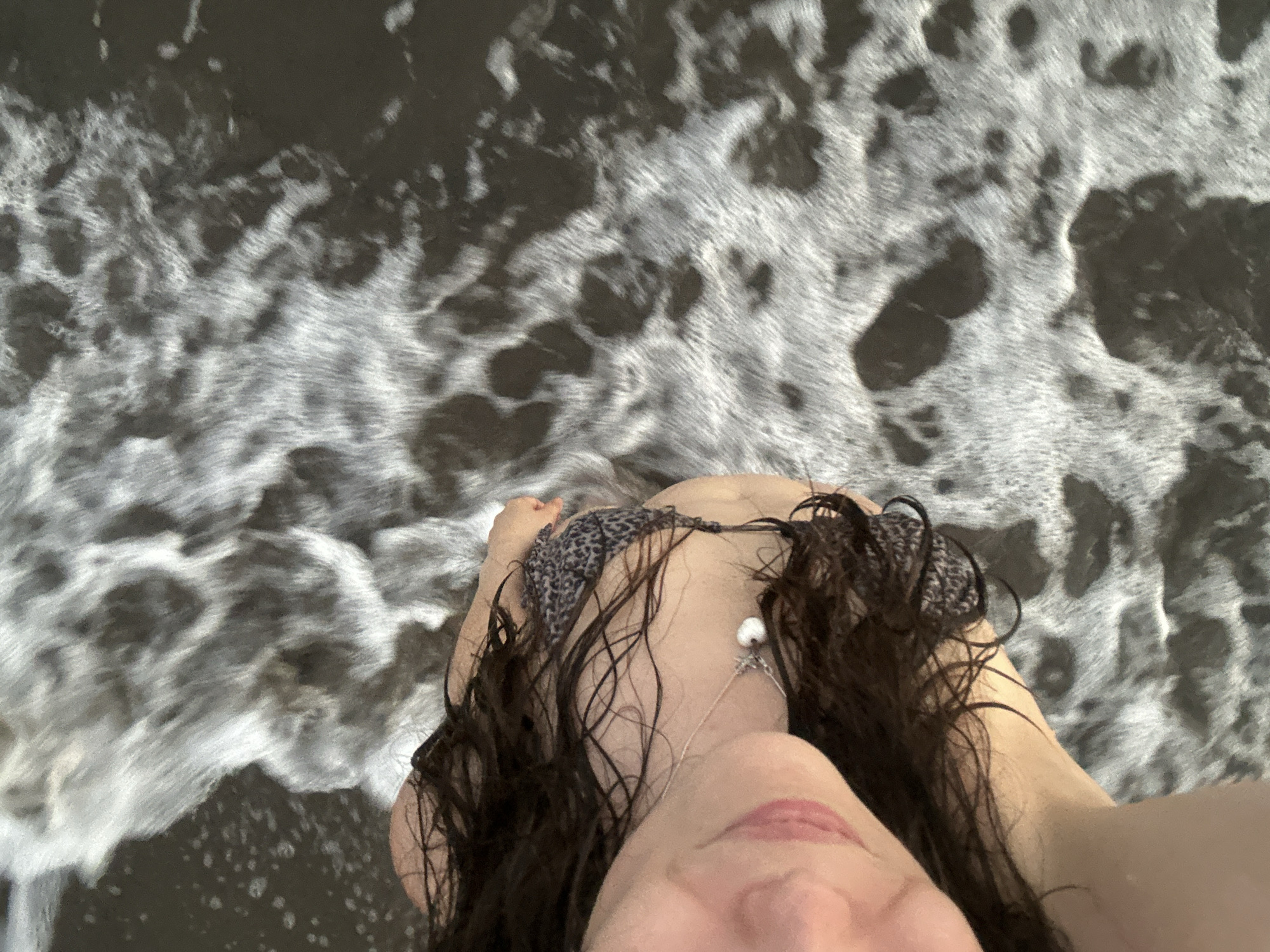 A close-up, overhead shot captures a person's head and shoulder as they lie on a furry, grey-and-white patterned surface. Their long, dark hair is spread out, and they are wearing a leopard-print bikini top. A delicate necklace with a white pearl charm is visible around their neck. The background is a dark, murky body of water with white foam and ripples, suggesting a beach or coastal area. There is no specific indication of the city of Ortaca, Turkey, in the image itself.