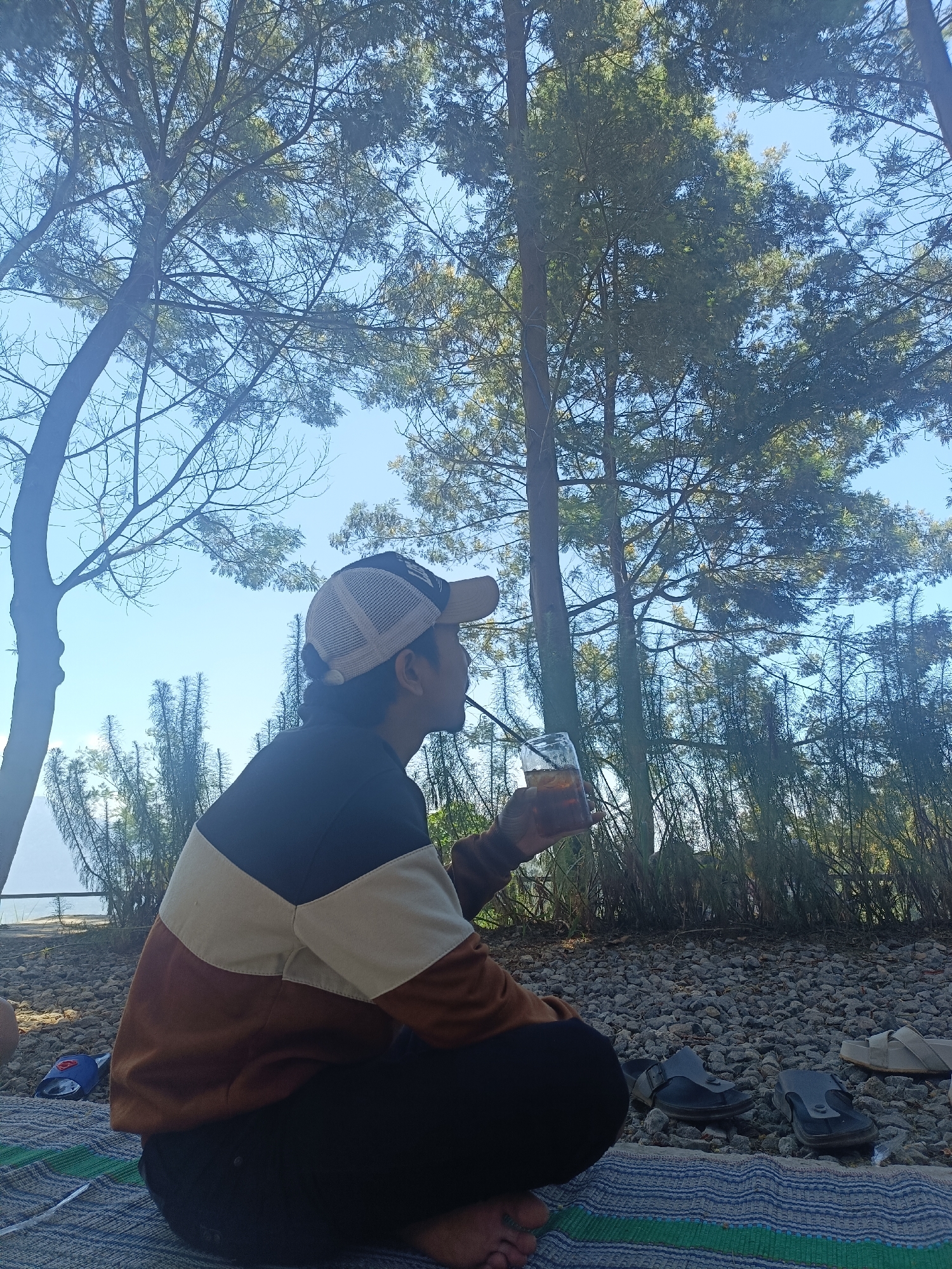 The image shows a person sitting on a mat outdoors, holding a glass of iced tea. The person is wearing a brown, beige, and black color-blocked sweatshirt and a baseball cap. The background consists of trees and a rocky ground. The scene appears to be set in a natural area, possibly a park or forest, in Kopeng, Indonesia. The lighting suggests it is daytime. There are several pairs of sandals visible on the ground near the person.