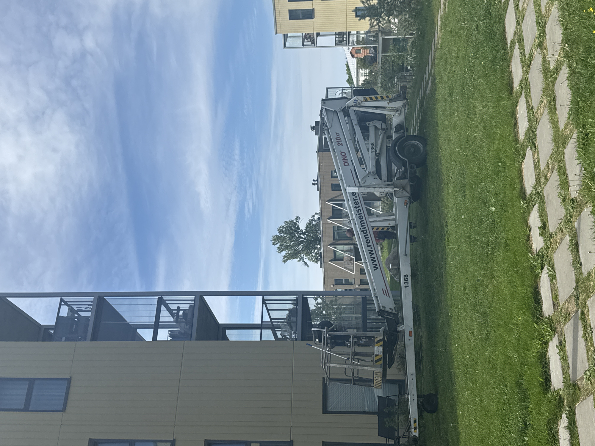 A white trailer-mounted aerial work platform, identified by the brand name "DINO 210 xt" and the number "1368", is parked on a grassy area in front of modern residential buildings in Tallinn, Estonia. The platform has a person in its basket, appearing to be working. The buildings, characterized by light-colored facades and balconies with glass railings, are situated on the left and right sides of the frame. The sky is partly cloudy with a blue hue. A path made of square concrete pavers is visible in the foreground.