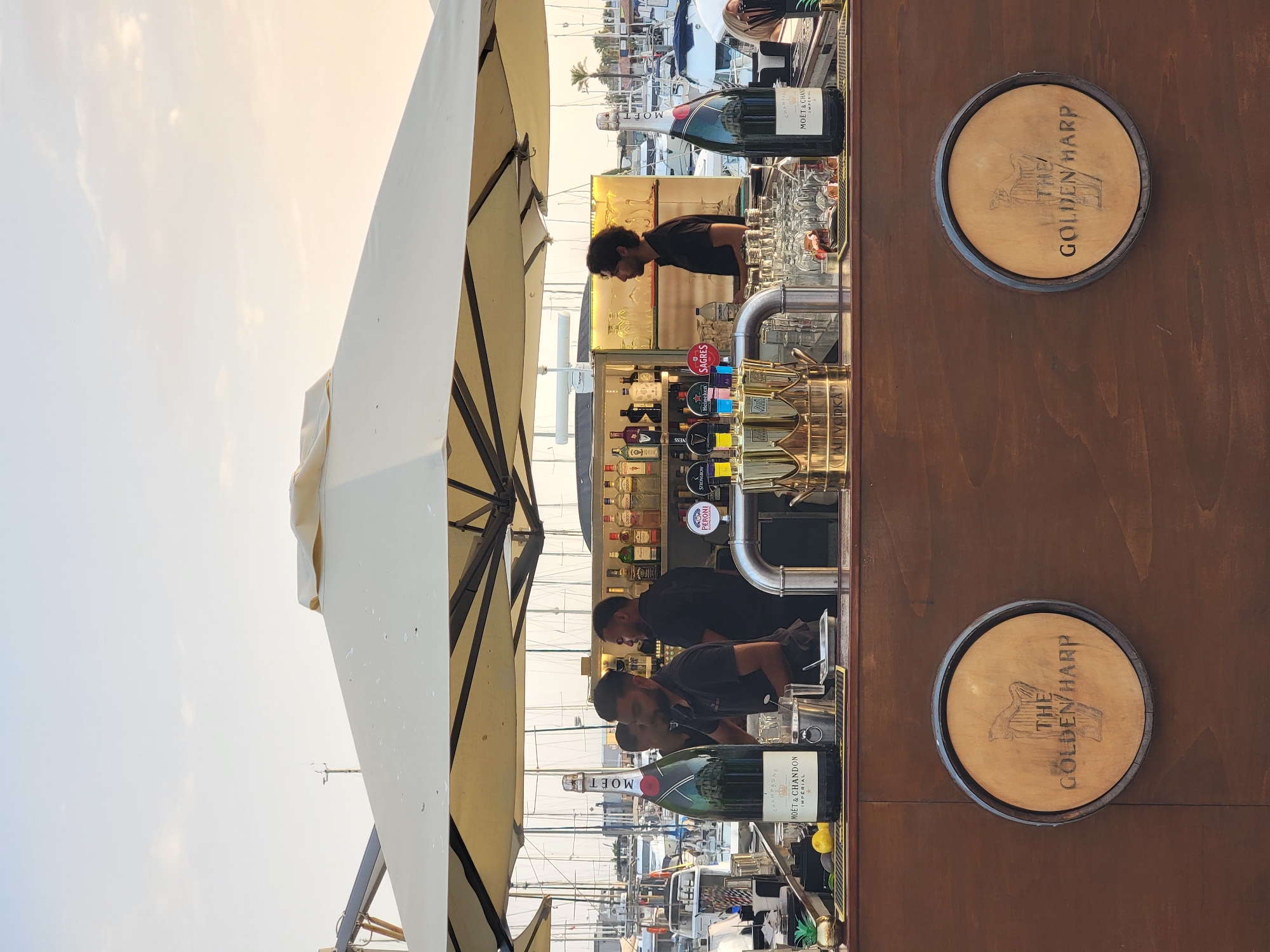 The image depicts an outdoor bar scene at a marina, likely in Quarteira, Portugal, given the context provided. The foreground features a wooden bar counter with two circular wooden emblems displaying a harp and the text "THE GOLDEN HARP." On the bar, there is a large bottle of Moët & Chandon Champagne, several other bottles of alcohol, glassware, and bar tools. Three men, presumably bartenders, are visible behind the counter. One man in the center is interacting with the bar equipment, while two others stand to his left and right. The background shows a bright, overcast sky and numerous sailboats docked in the marina. The overall atmosphere is casual and suggestive of a social gathering or establishment.