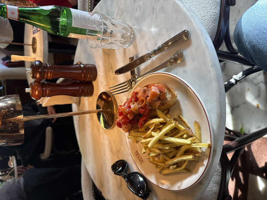 This image depicts a close-up, first-person perspective of a meal served at Caffè Svizzero in Piazza Costituzione, Cagliari, Italy. The central object is a round, white marble table. On the table, a white ceramic plate with a red rim displays a portion of golden-brown french fries and two pieces of bruschetta, each topped with diced red tomatoes. The plate is inscribed with "SVIZZERO." To the left of the plate, a pair of black sunglasses rests on the table surface. To the right of the plate, a silver fork and a silver knife, featuring an ornate handle, are placed. The knife rests on a small, metallic knife rest. An empty, clear, ribbed glass stands to the right of the cutlery, next to a partially visible green glass bottle of mineral water. Behind the plate, two dark brown wooden shakers, likely for salt and pepper, are present. A small, brass-colored table lamp with an inverted cup-shaped shade is positioned behind the food.
The background is out of focus, revealing a restaurant interior with other tables and blurred figures. A person wearing a dark top is partially visible to the left of the lamp. Another individual in a white shirt is discernible behind the shakers. A hand with a red sleeve is visible to the far right, near the water bottle. The lower right corner of the image shows the knee and thigh of a person wearing light blue jeans, indicating the photographer's seated position at the table. The flooring beneath the table appears to be a light-colored tile or marble with dark patterns.