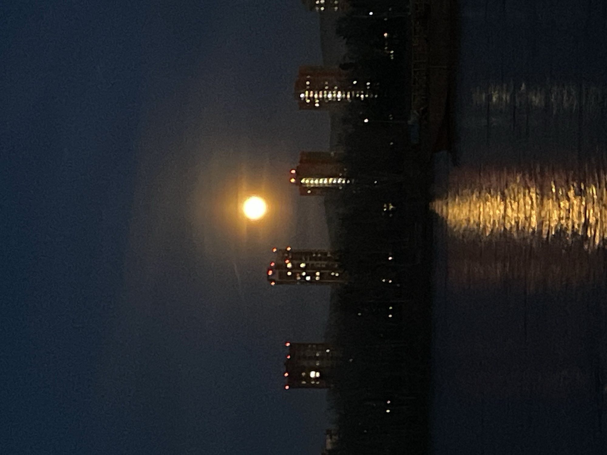 The image displays a nocturnal urban landscape, likely in Krasnoyarsk, Russia, as suggested by the location context. A prominent, bright, and luminous full moon hangs in a dark, clear sky, dominating the upper central portion of the scene.
To the right of the moon, several tall buildings are silhouetted against the night sky. These structures appear to be multi-story, possibly residential or office buildings, with numerous windows emitting warm, yellow light. Some of these buildings also feature small, red lights near their tops, which could be aircraft warning lights.
Below the buildings, a body of water, likely a river, stretches across the lower part of the frame. The surface of the water intensely reflects the artificial lights from the buildings, creating shimmering, elongated vertical streaks of golden-yellow light. These reflections suggest a gentle rippling or movement on the water's surface.
The overall scene conveys a peaceful and atmospheric ambiance, characteristic of a city night under a bright moon. There are no people visible, and no specific activity or event is taking place beyond the natural occurrence of moonlight and city illumination. No visible text is present in the image.