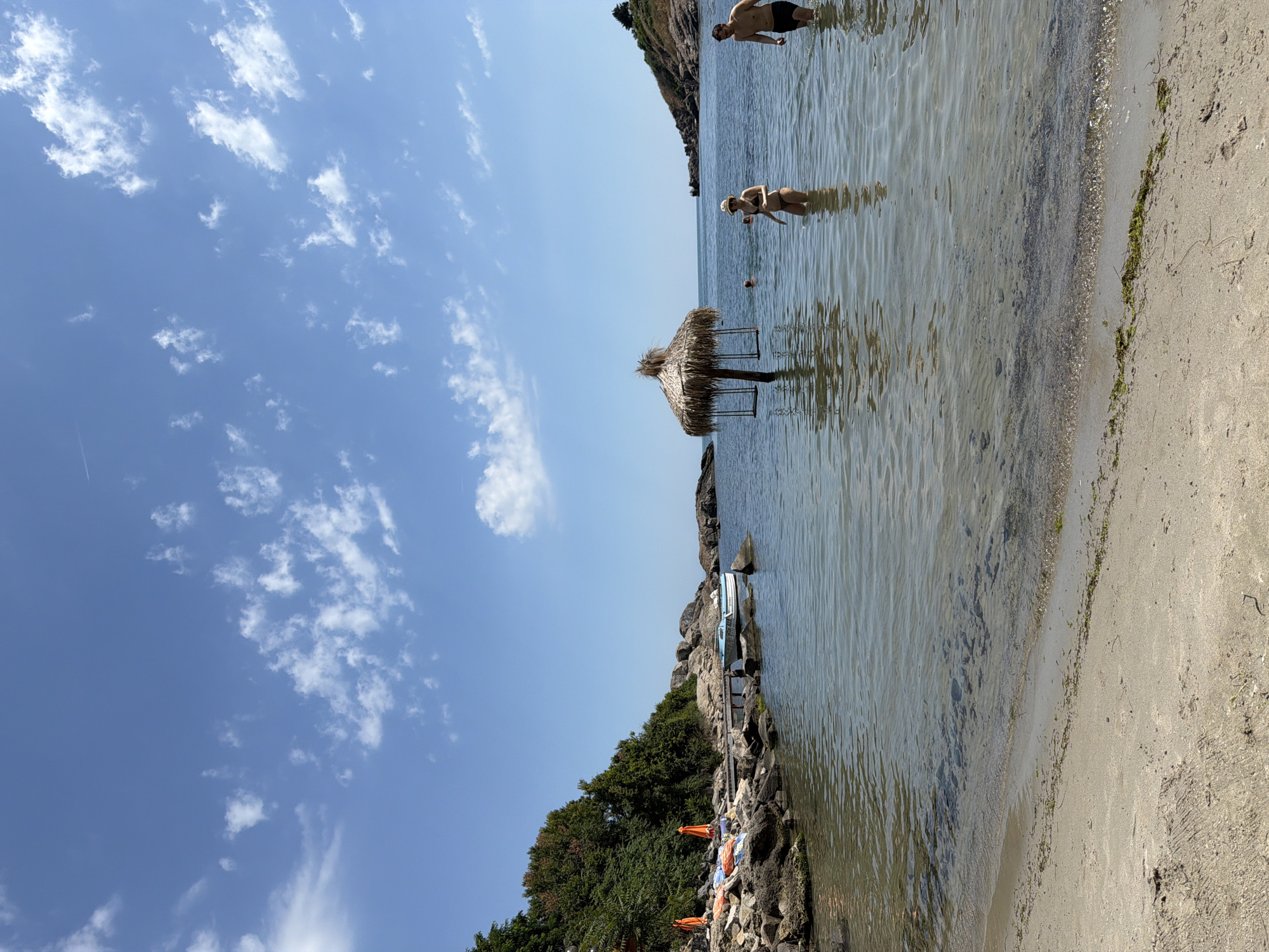 The image depicts a beach scene in Sozopol, Bulgaria, under a clear blue sky with scattered white clouds. The orientation of the photo is sideways. In the foreground, the sandy beach meets the calm, shallow water. The water is clear, allowing visibility of the sandy bottom.
Several people are visible in the water. A man in black swim trunks stands further out in the water, while a woman wearing a bikini and a hat is closer to the shore. There are also red buoys in the water, suggesting a designated swimming area.
A straw-roofed umbrella, supported by poles, stands in the water near the shore. Further in the background, rocky cliffs covered with dense green trees rise from the water. On the rocky shoreline, several beach chairs and orange umbrellas are visible, along with a boat docked near the shore.
The time of day appears to be daytime, with bright sunlight casting reflections on the water's surface. The weather seems warm and pleasant, suitable for a beach outing. The overall atmosphere is relaxed and leisurely, indicative of a summer day at the seaside. No visible text is present in the image.