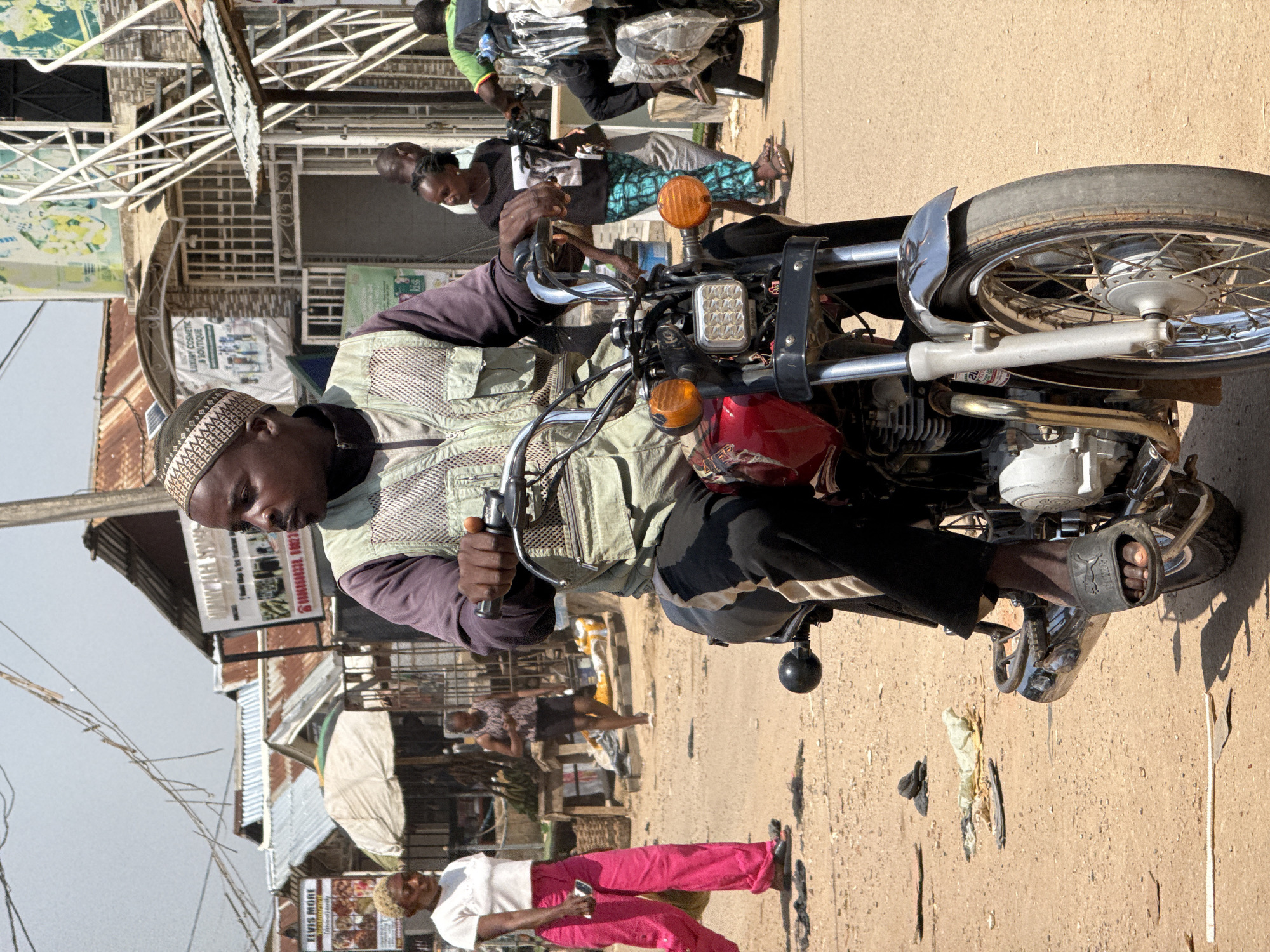 The image captures a street scene in Abuja, Nigeria, with a man riding a motorcycle as the primary subject. He is wearing a light-colored vest over a dark shirt and a patterned cap. His right hand is on the motorcycle's handlebars, and his left hand appears to be holding something. The motorcycle itself is red and silver, with a prominent chrome exhaust pipe and a visible engine. The background shows a bustling street environment with various buildings, some with corrugated iron roofs, suggesting a commercial or residential area. Other people are visible in the background, some walking and others on motorcycles, indicating everyday activity.
The time of day appears to be daytime, with clear sunlight casting shadows on the dusty ground. The weather seems dry and warm, typical of a sunny day in Abuja. Visible text includes "LUXURY COSMETIC & BOUTIQUE" and what appears to be a phone number on a sign in the background, adding to the urban context. Another sign reads "ELVIS MORE (Physical Body)". The overall mood is one of daily life and commerce in a vibrant street setting. The man on the motorcycle appears focused on his ride.