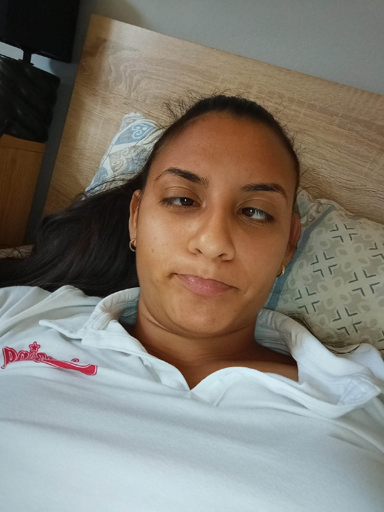 A woman lies on a bed, with her head resting on a pillow. She is wearing a white polo shirt with a red logo. The room appears to be a bedroom, with a wooden headboard and a grey wall. There is a dark object, possibly a television, visible on the left side of the image. The woman's hair is long and dark, and she has an earring in her left ear. The image is a close-up shot, focusing on the woman's face and upper body. The scene is indoors and appears to be in a private residence. The location is not specified as being in the city of Yambol, Bulgaria.