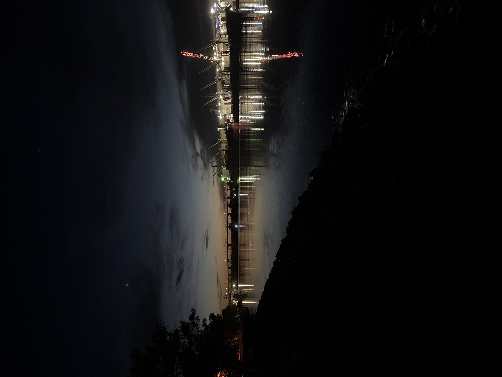 This nighttime image depicts the Port of Varna in Varna, Bulgaria. The port's cranes and structures are illuminated, creating a reflection on the water's surface. The sky shows a gradient from dark to twilight, with clouds present. The foreground features dark landmass with hints of vegetation.