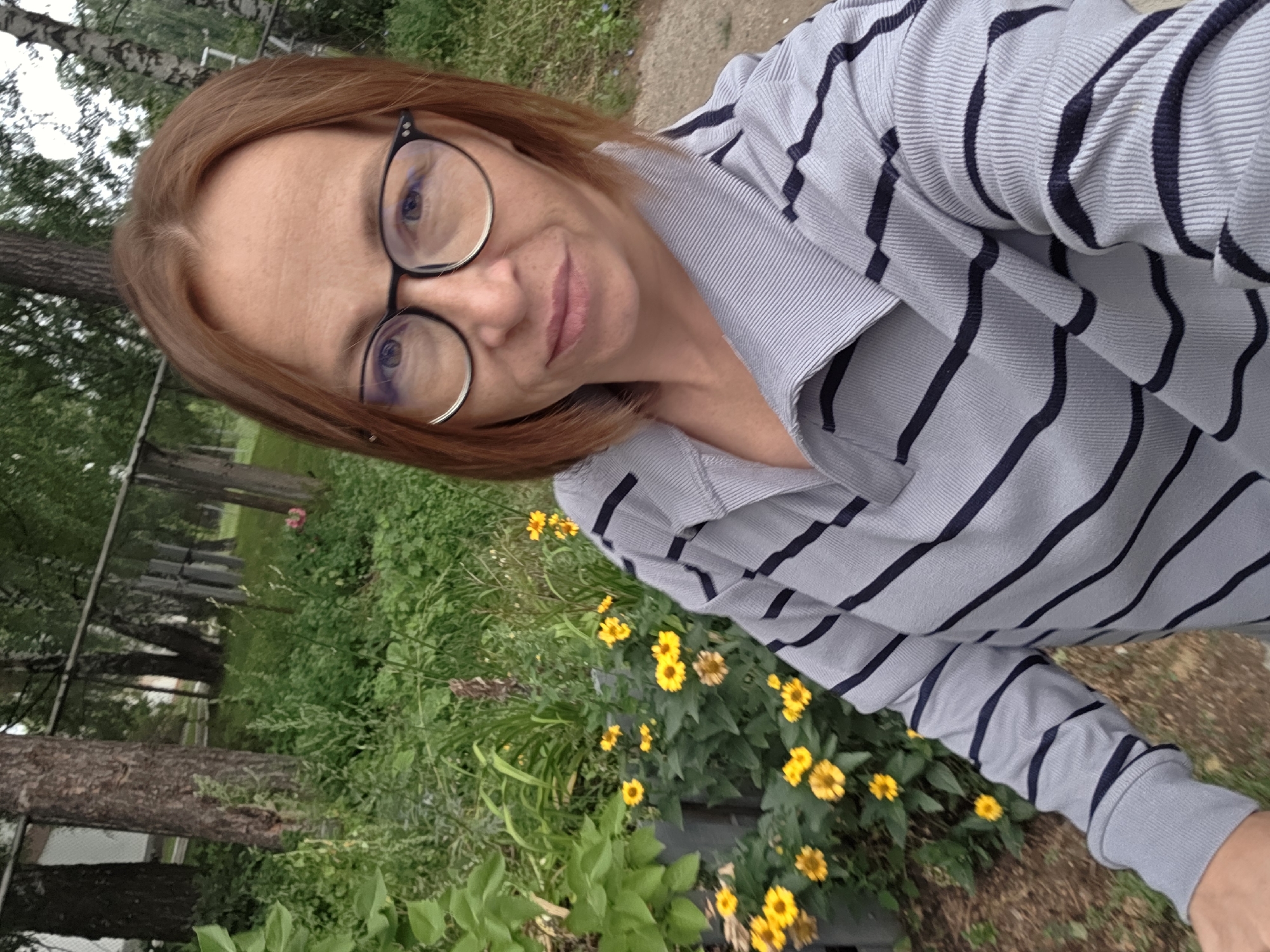 In a self-portrait taken outdoors, a woman with reddish-brown hair and glasses is positioned in the foreground, looking at the camera. She is wearing a grey, long-sleeved, collared shirt with dark navy stripes. Behind her, a garden area is visible with numerous yellow flowers and green foliage. In the background, a chain-link fence runs horizontally across the image, with trees and greenery beyond. The scene appears to be in a park or residential area, possibly in Chaikovsky, Russia, given the context provided.