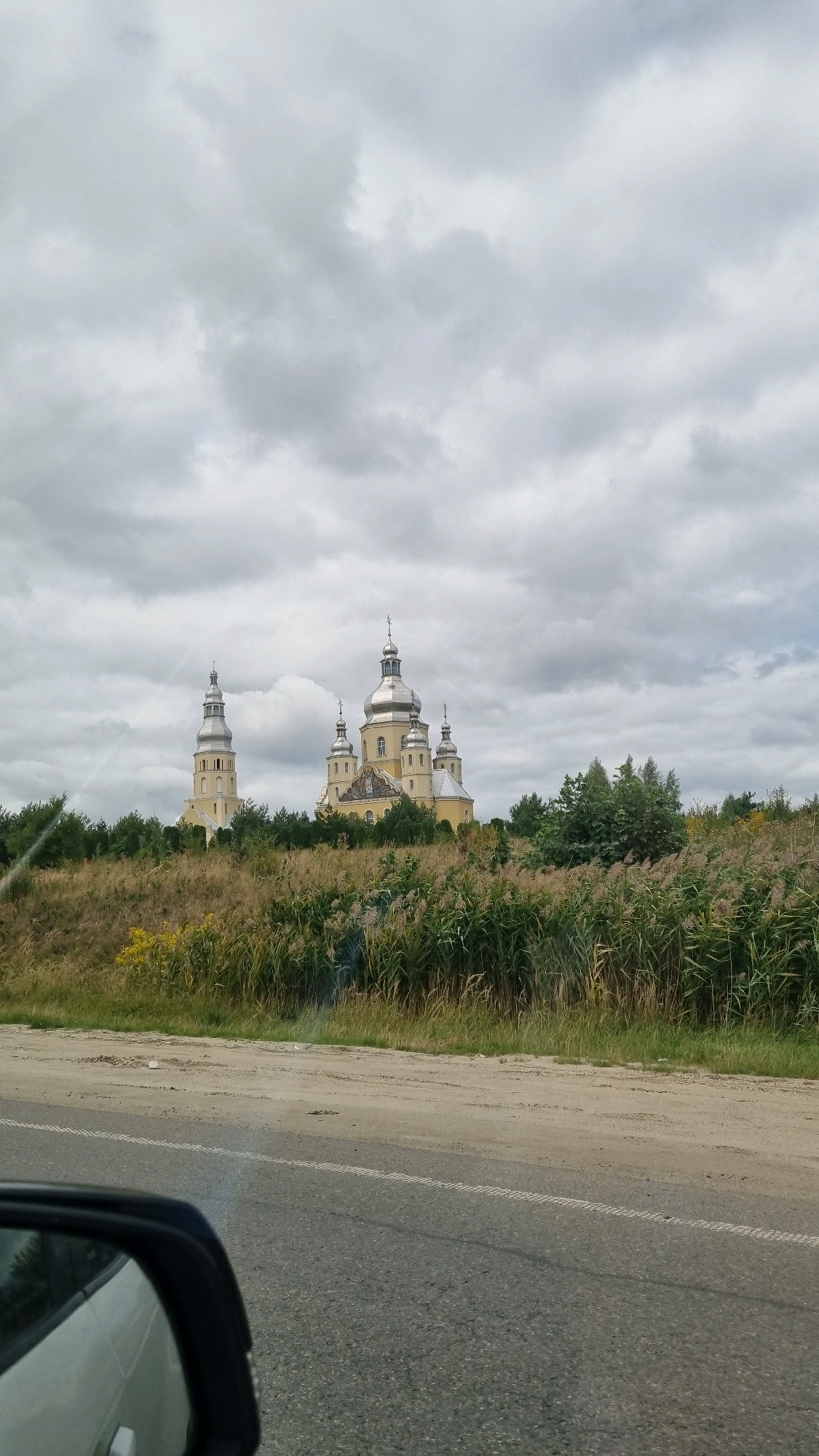 The image shows the Church of the Transfiguration in Zubra, Ukraine, under a cloudy sky. The yellow church, with its silver domes and a tall bell tower, is situated on a hill surrounded by greenery and tall reeds. The foreground features a road with a white line marking, and a car's side mirror is visible on the left. The scene appears to be captured from a moving vehicle.