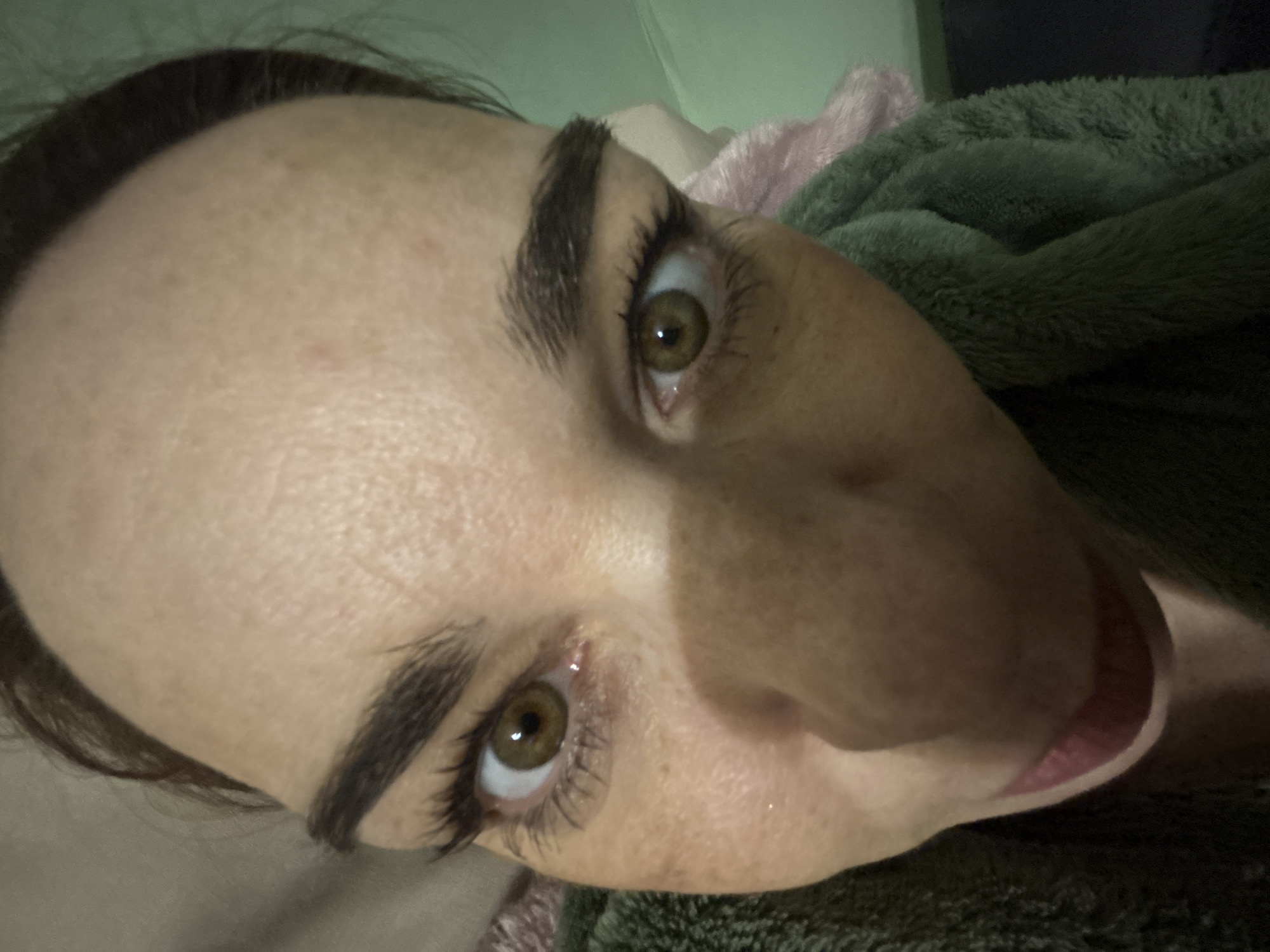 A close-up, tilted view of a person's face, likely a selfie, with the subject lying down. The person has dark hair, prominent eyebrows, and hazel-colored eyes. A portion of a green and pink blanket or bedding is visible in the background. The environment appears to be indoors, possibly a bedroom. There is no information available in the image to determine the location as Vsevolozhsk, Russia.