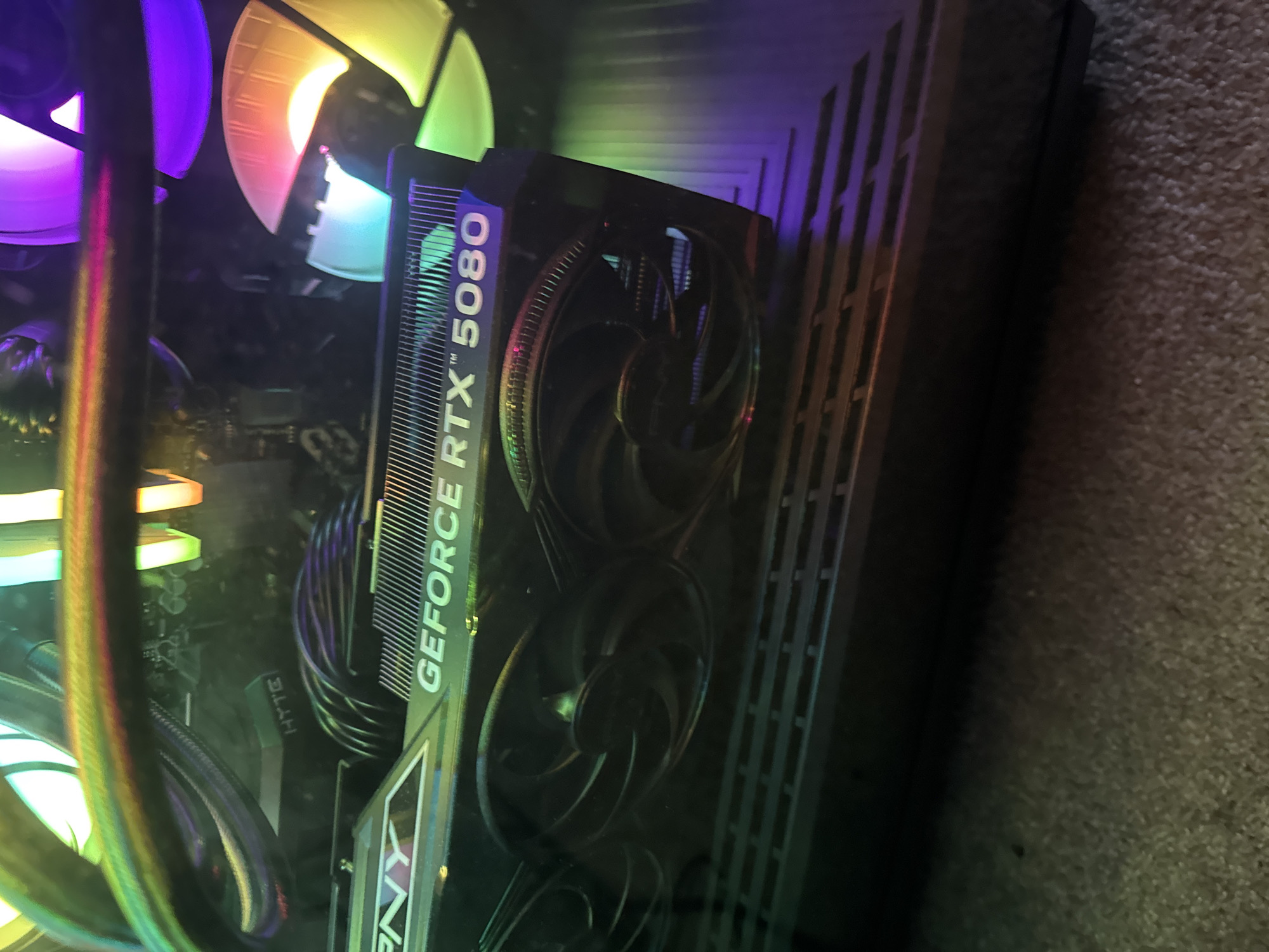 The image shows a close-up view inside a computer case. The dominant feature is a PNY GeForce RTX 5080 graphics card. To the left are RGB illuminated cooling fans and liquid cooling tubes. The computer case is dark. The background shows a section of carpet. The location in the city of Glendale Heights, United States of America cannot be determined from this image.