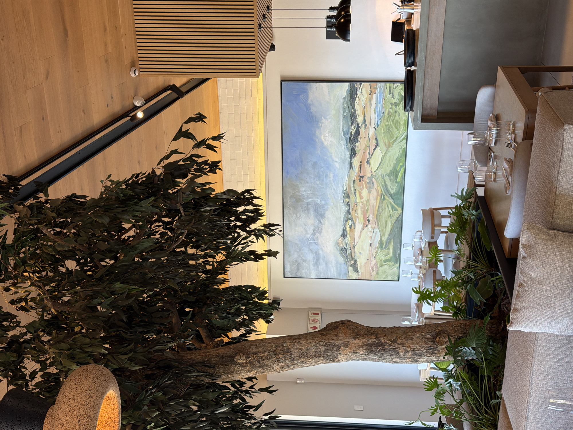 The image, oriented vertically, showcases a modern and elegantly designed indoor space, likely a restaurant or hotel lounge. No people are present.
The scene is dominated by a large, realistic artificial tree with a prominent, gnarled trunk and dense green foliage, extending from the bottom left and across the center of the frame. Its branches create a natural division within the expansive room.
To the right of the tree, a sophisticated dining area is arranged with multiple tables, each meticulously set with clear glasses, plates, and cutlery. Seating consists of comfortable, upholstered chairs and bench-style seating in neutral, earthy tones. Behind these tables, a long, light gray counter or bar area is visible, with more glassware and serving dishes stacked.
A striking feature on the main wall is a large, horizontal landscape painting. It depicts a scenic vista with a blend of light blues, greens, and ochre, executed with broad, impressionistic brushstrokes. Below the painting, on a white panel near the tree trunk, a set of red directional icons is visible: two arrows (one pointing right, one up) and a circular icon.
The room's aesthetic is contemporary, utilizing a palette of light woods for flooring and ceiling slats, white and off-white walls, and track lighting with black fixtures. An illuminated LED strip runs along a white brick accent wall, adding to the ambient light. On the wooden ceiling, a white circular smoke detector branded "KIDDE" and another "System Sensor" device are visible. The overall impression is one of a spacious, well-lit, and refined environment, suggesting readiness for guests during daytime hours. The location is Drakenstein Nu, South Africa.