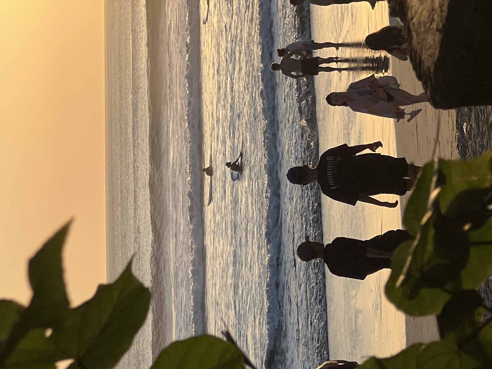 In Kuta, Indonesia, during sunset, the scene unfolds on a beach with people scattered throughout. The sun, low in the sky, casts a warm, orange glow over the ocean, its light reflecting off the water and creating shimmering waves that approach the shore. Several individuals are in the ocean, with at least three on surfboards, one actively paddling. On the wet sand near the waterline, a group of people are visible. Two young men in dark t-shirts stand facing the ocean, while further down the beach, a couple stands together. Another young woman walks along the shore, carrying a towel and holding flip-flops, with a woman sitting nearby. In the foreground, out of focus, are large green leaves and a dark, textured surface, suggesting they are part of a natural or built structure at the edge of the beach. The overall atmosphere is serene, capturing a typical evening at a popular beach destination.